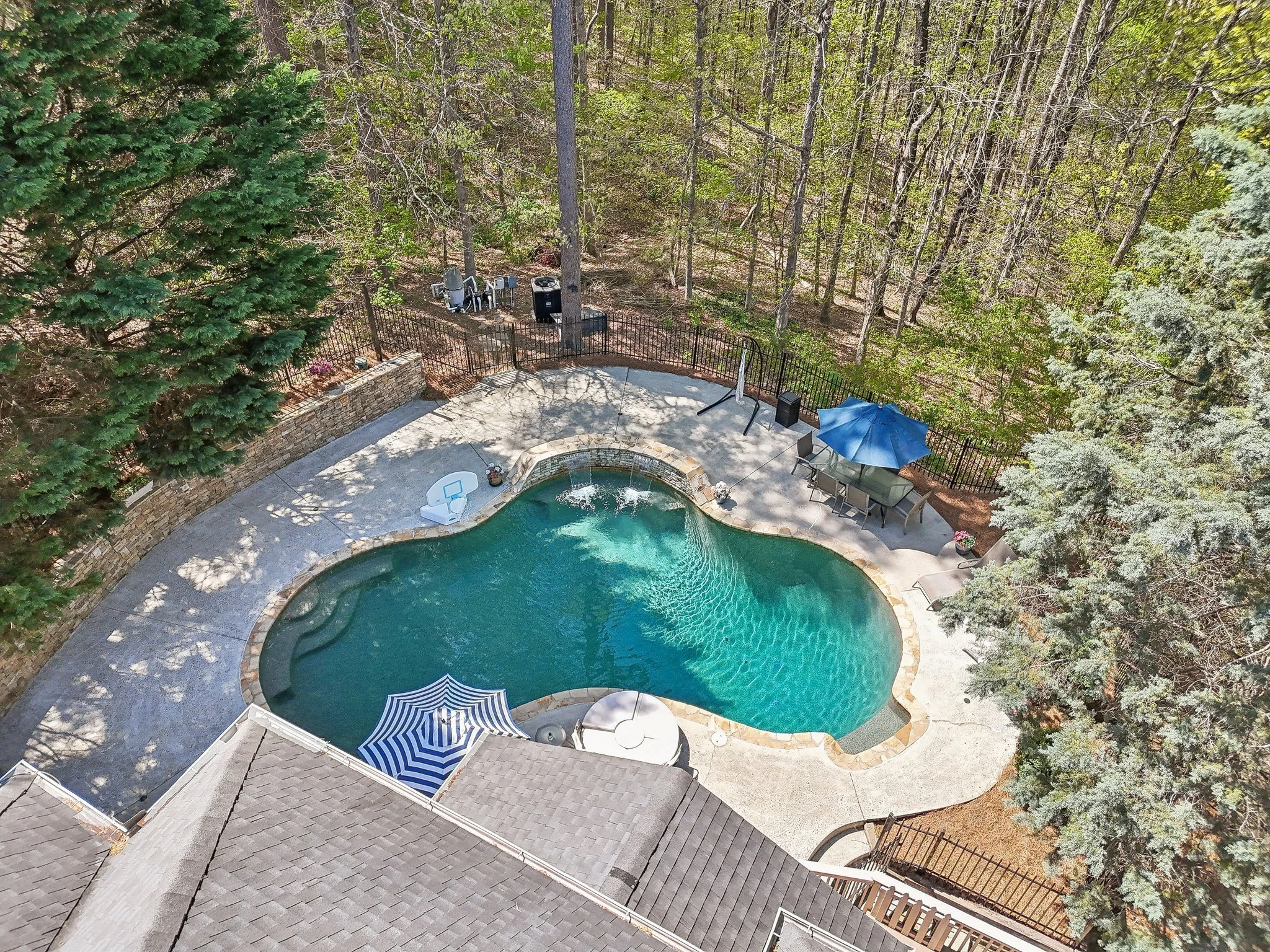 Aerial view of a backyard swimming pool surrounded by concrete decking and trees. There are patio chairs, a table with a blue umbrella, and a striped pool float in the water. A rock wall borders part of the area.