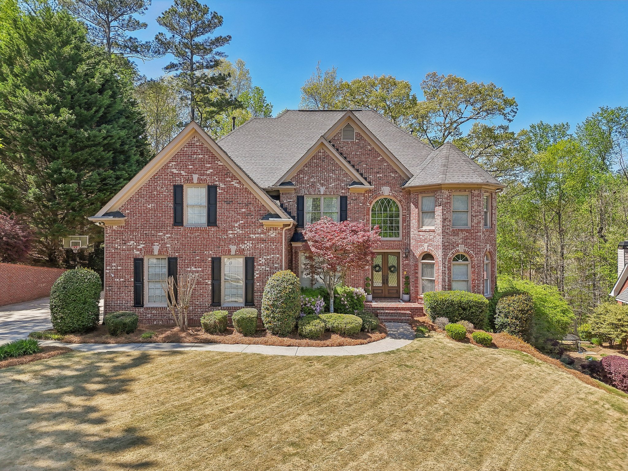 Two-story red brick house with black shutters, surrounded by a manicured lawn and landscaping, featuring trees and bushes. A basketball hoop is visible to the left.