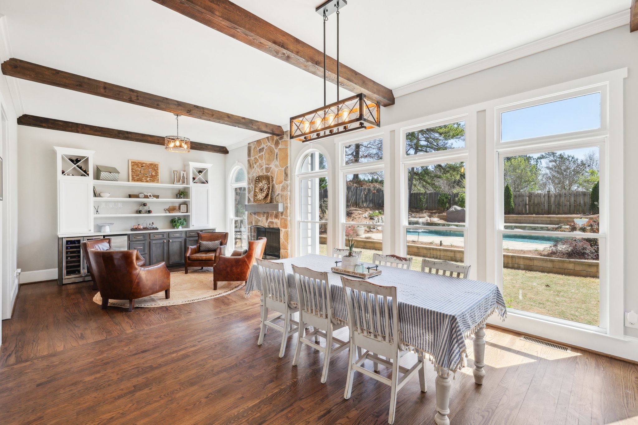 Spacious living and dining area with wooden beams, a stone fireplace, leather chairs, built-in shelves, and a dining table with chairs in front of large windows with a view of the backyard and pool.