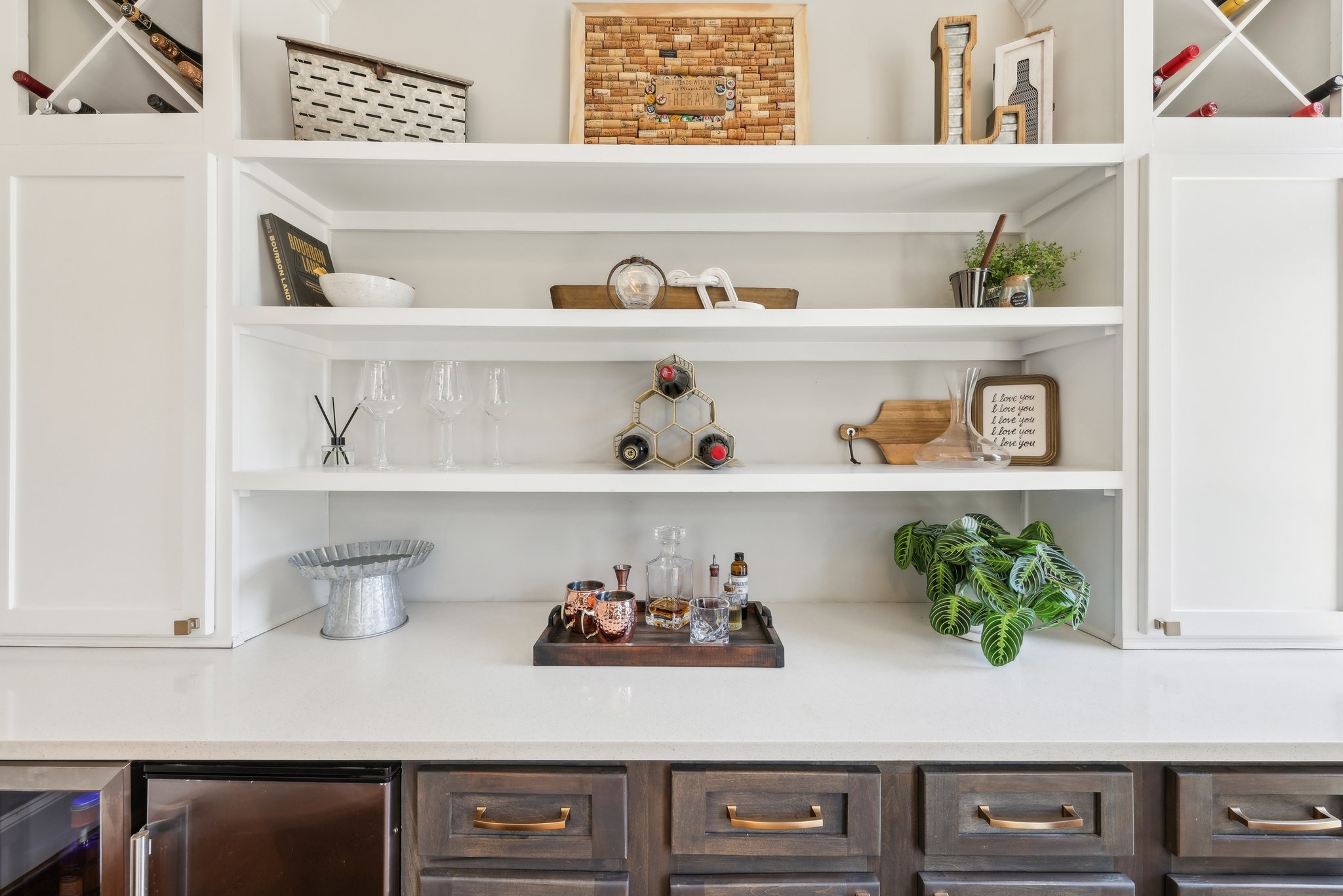 White kitchen shelves with decorative items, glassware, and a small plant. A countertop features a tray with bar accessories, liquor bottles, and a metal stand.