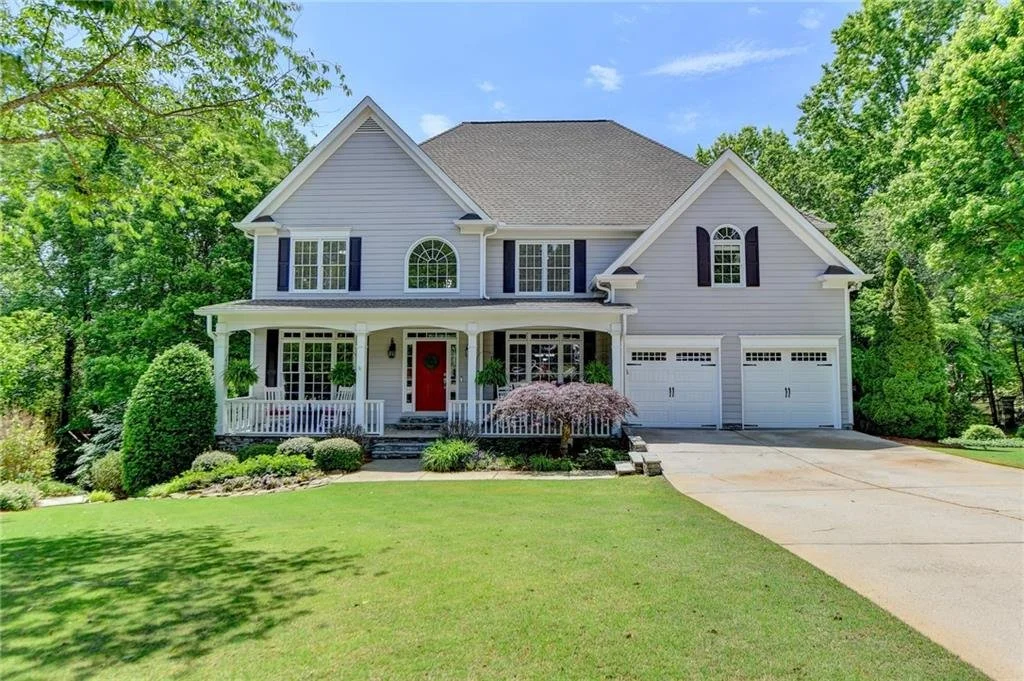 Two-story suburban house with gray siding, red door, black shutters, and two-car garage, surrounded by green lawn and trees.