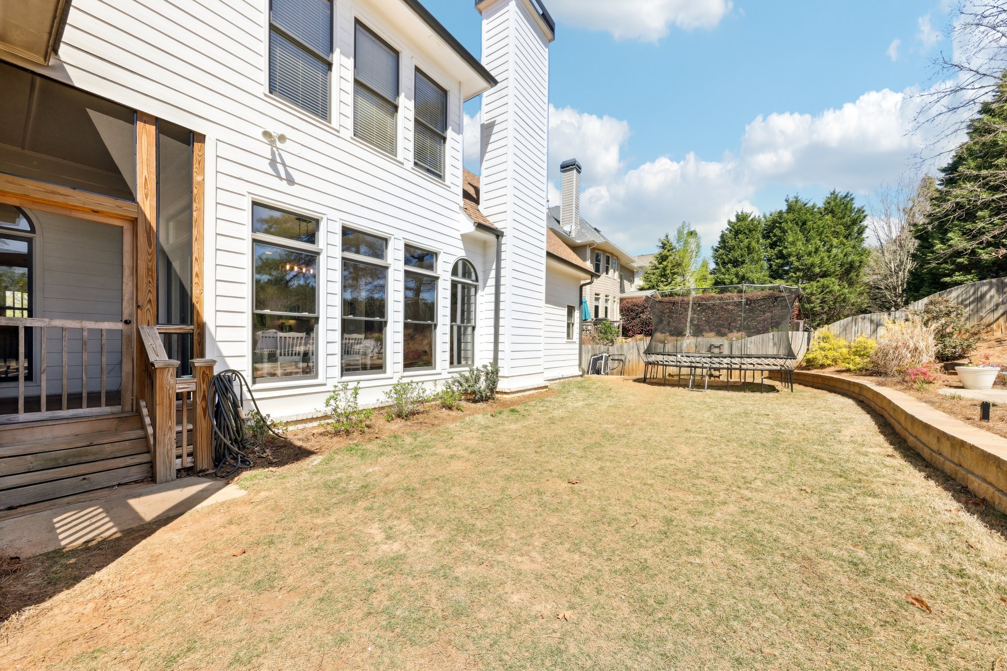 Backyard of a house with white siding, featuring a wooden porch, large windows, a trampoline, grass, and a retaining wall with plants, under a blue sky with fluffy clouds.