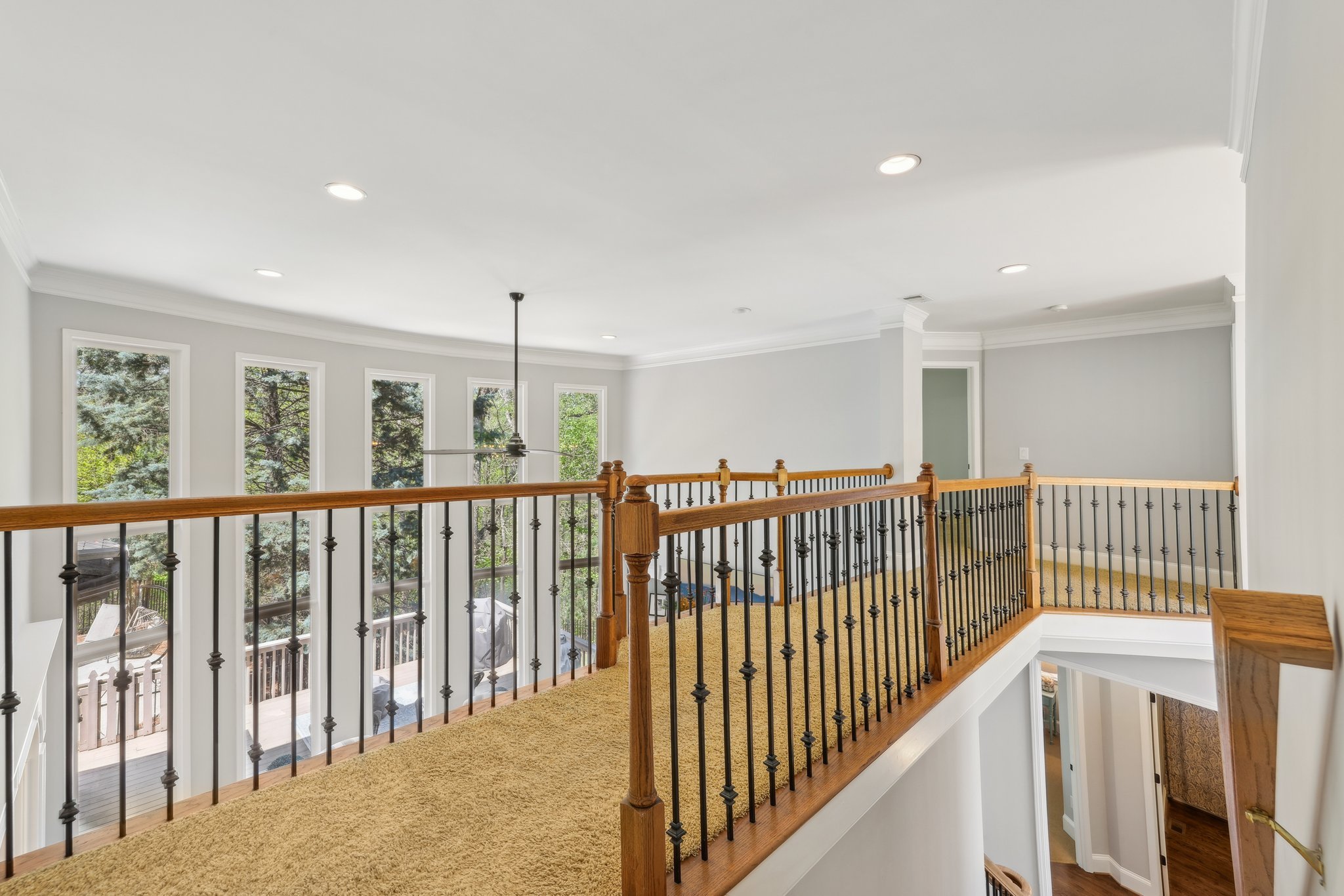 Interior of a modern home with a carpeted upstairs hallway. Features include a wooden railing with black balusters, recessed lighting, large windows providing natural light, and a view of the surrounding trees. A ceiling fan hangs from the ceiling. A