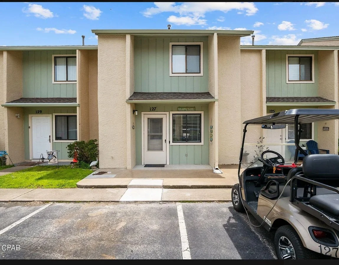Exterior of a townhouse with a beige and green facade, featuring three units labeled 125, 127, and 129. A golf cart is parked in front of unit 127 on a concrete driveway. The sky is blue with scattered clouds.