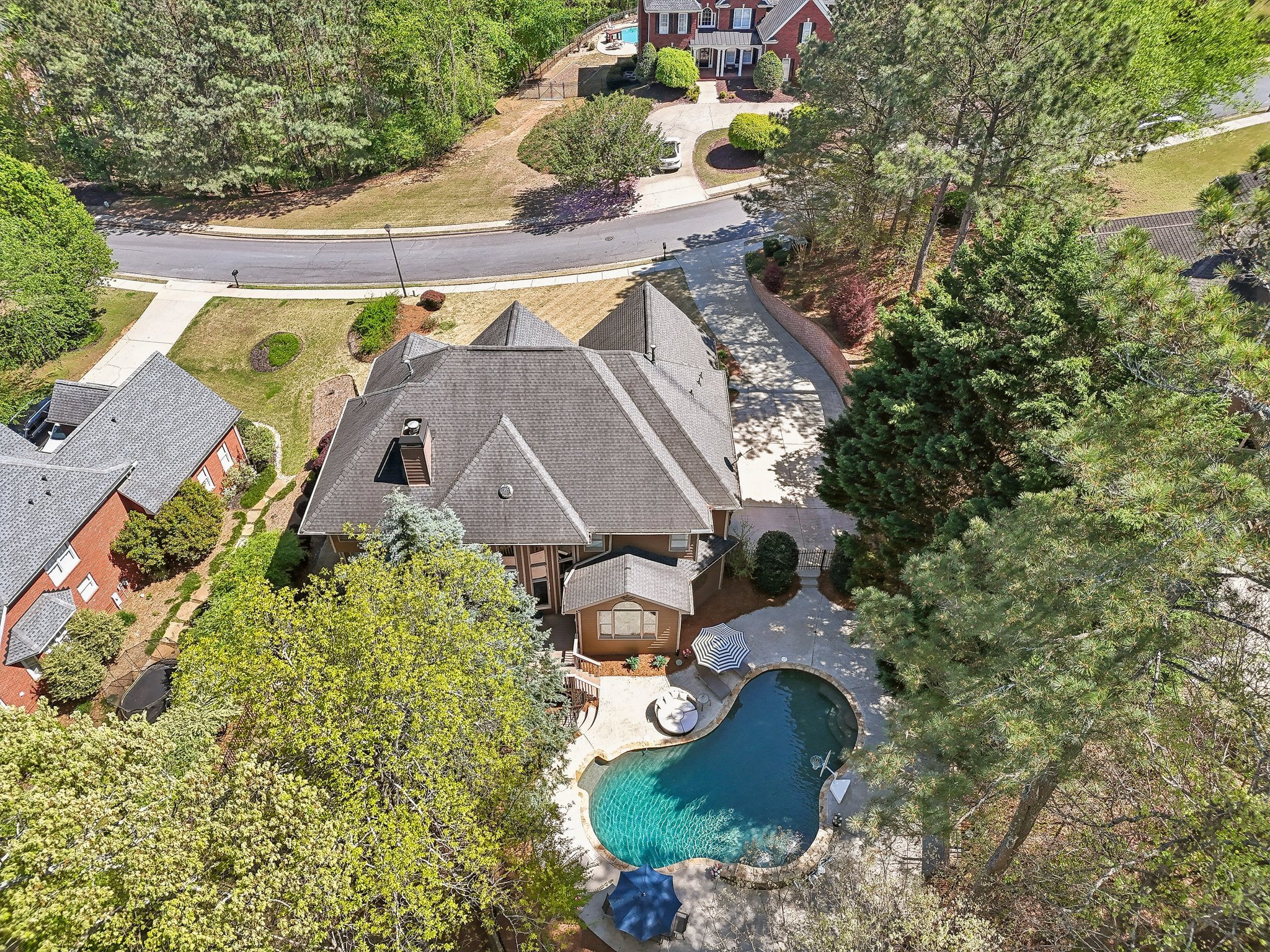Aerial view of a residential neighborhood with houses, trees, and a backyard with a swimming pool. The main house has a complex roof design and is surrounded by greenery and a paved driveway.