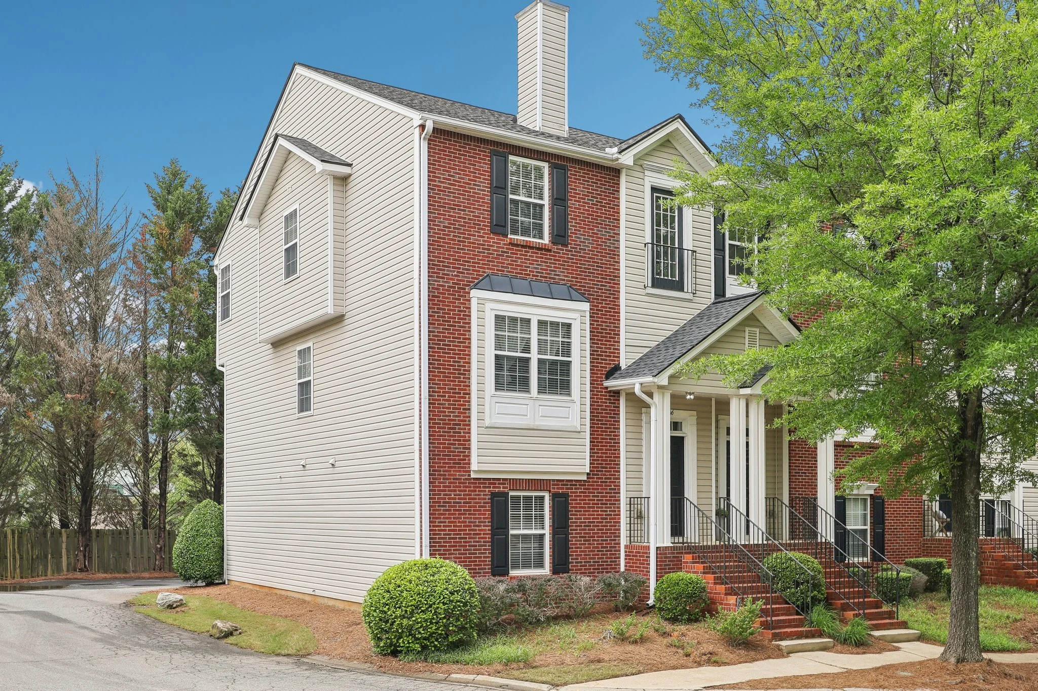 A multi-story residential building with a combination of red brick and white siding exterior, black window shutters, and a small front porch with brick stairs and black railings. There are shrubs, trees, and a sidewalk in front, under a blue sky.