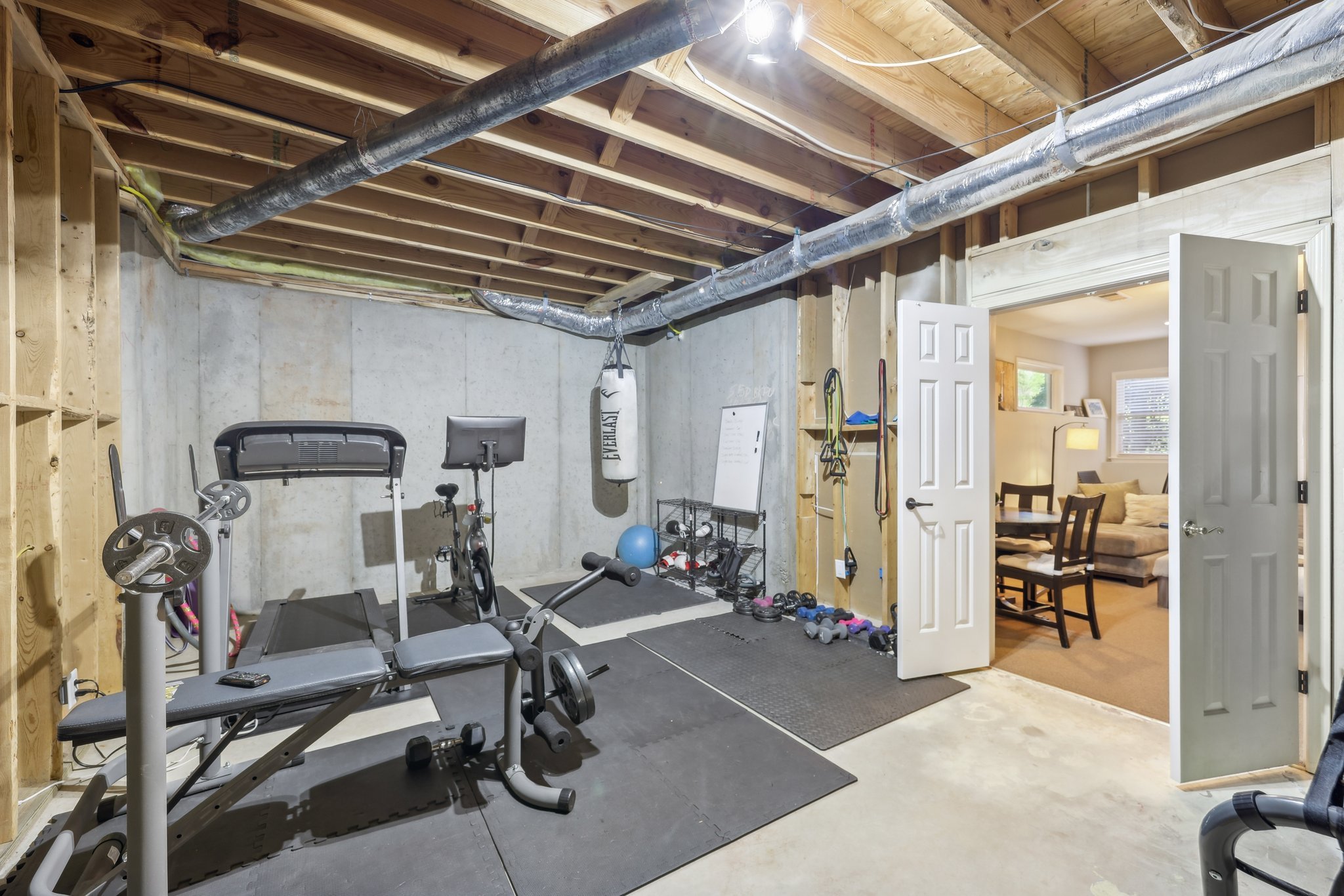A home gym with exercise equipment including a treadmill, stationary bike, bench press, and punching bag. Weights and resistance bands are on the floor. The room has exposed beams and ductwork, with an open door leading to a living area with a couch
