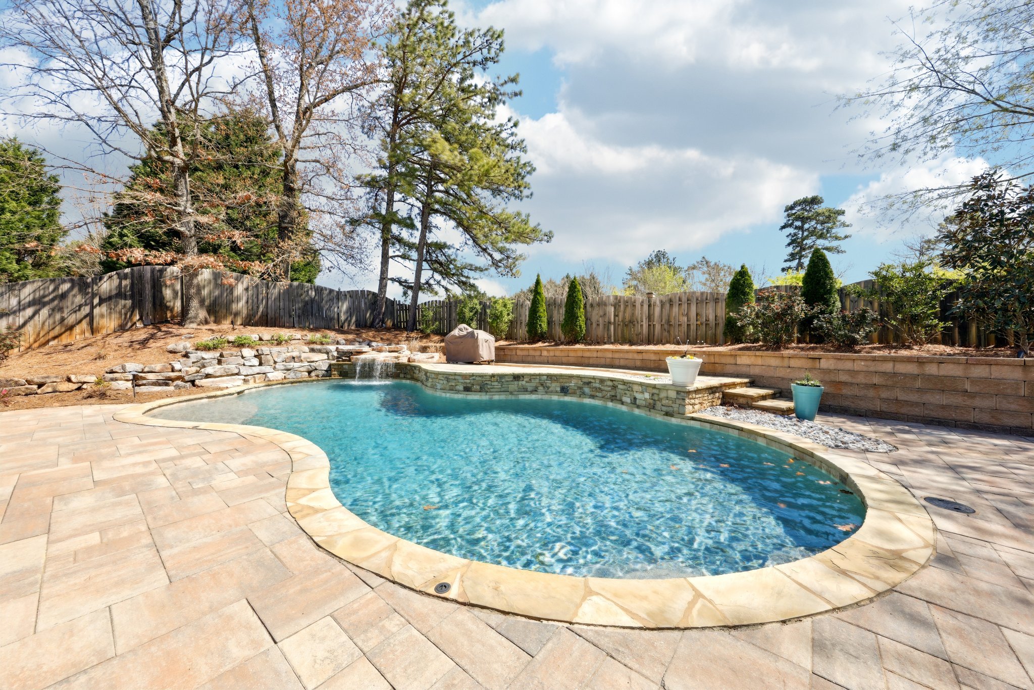 Backyard with a private swimming pool, stone patio, and surrounding wooden fence. Landscaping includes trees and shrubs.