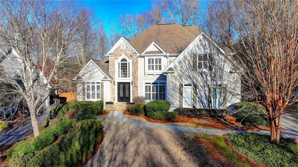 Two-story house with a stone facade, large windows, and dark front doors. Surrounded by a landscaped yard with shrubs and bare trees on a sunny day.