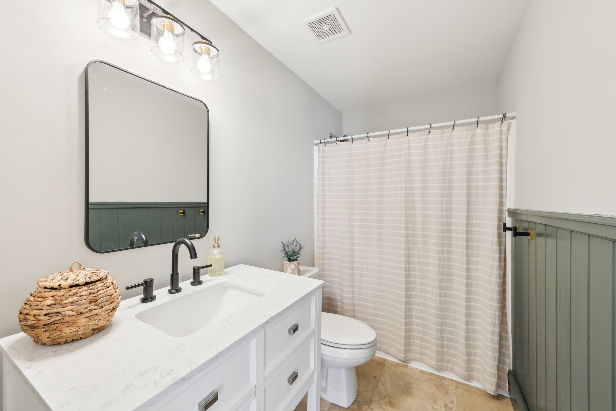 Modern bathroom with a white vanity and marble countertop, black faucet, wicker basket, lavender plant, large mirror, light fixtures, toilet, and shower with a striped curtain.