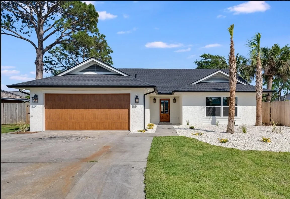Single-story house with white exterior, black roof, and wooden double garage doors. A concrete driveway, small front yard with grass, palm trees, and landscaping stones visible.