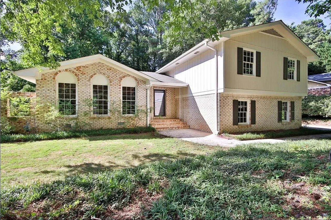 Exterior view of a two-story brick house with large windows, surrounded by a grassy lawn and trees.