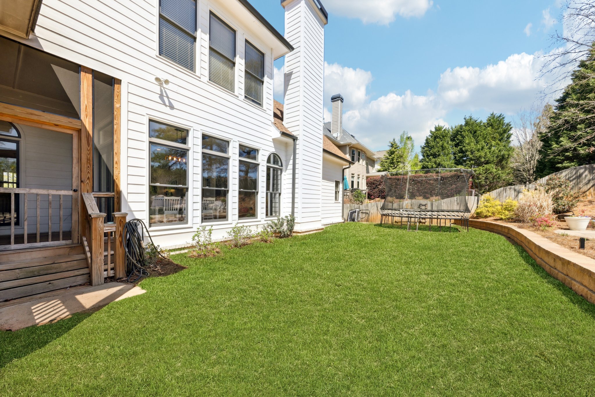Backyard with white house, large windows, green lawn, trampoline, wooden porch, and clear blue sky