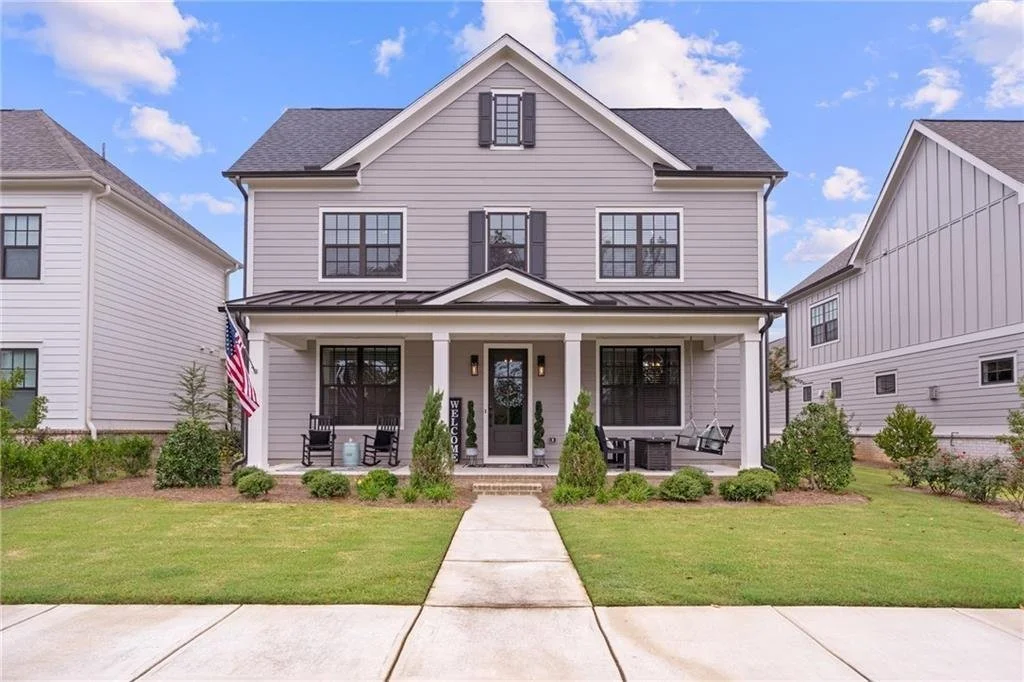 Front view of a modern two-story house with gray siding, black shutters, and a welcoming front porch. The porch features rocking chairs and a swing. An American flag is displayed near the entrance. Well-maintained lawn and shrubs are in the front yard.