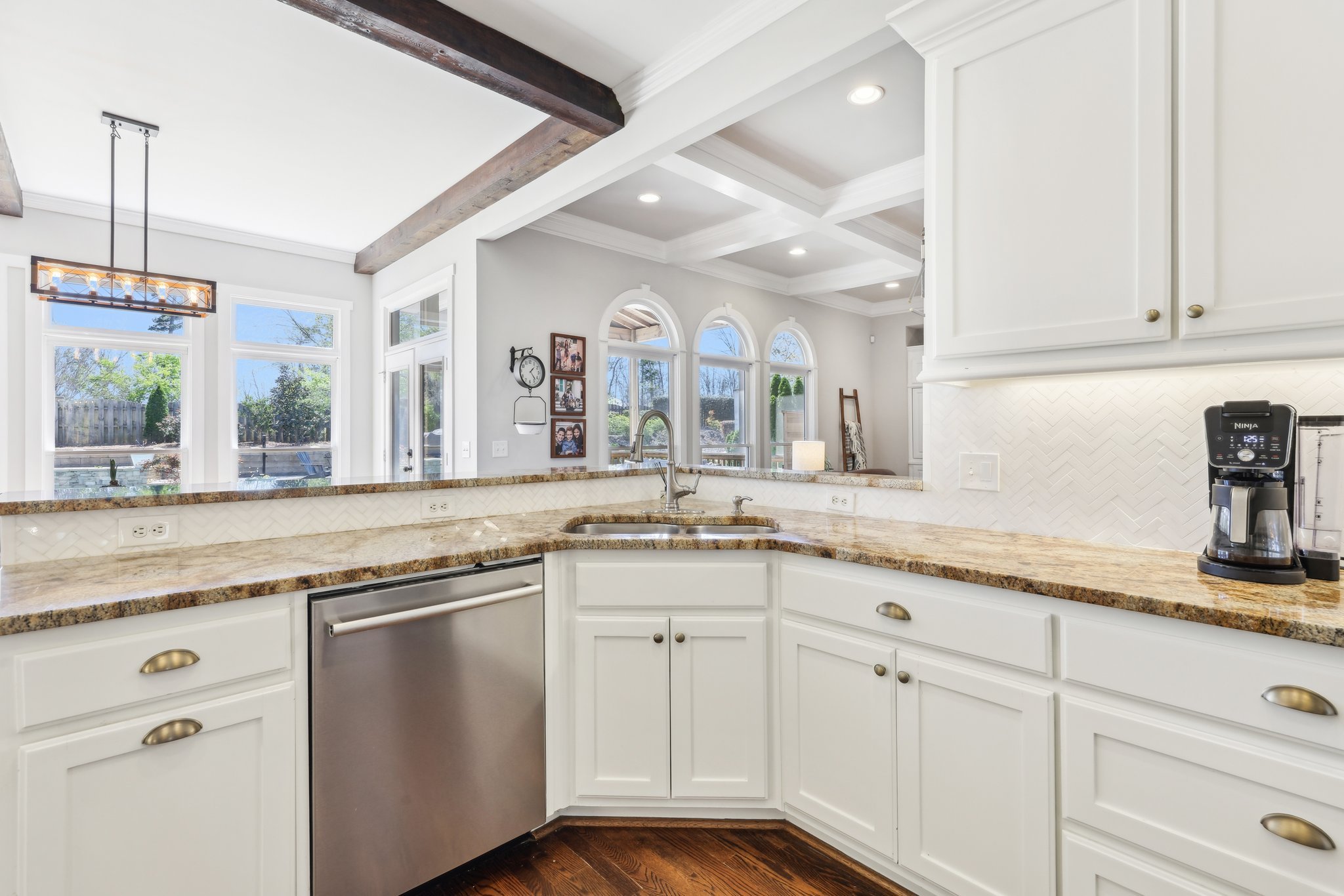 Modern kitchen with white cabinets, granite countertops, stainless steel dishwasher, coffee maker, and view of living area with large windows and wooden ceiling beams.