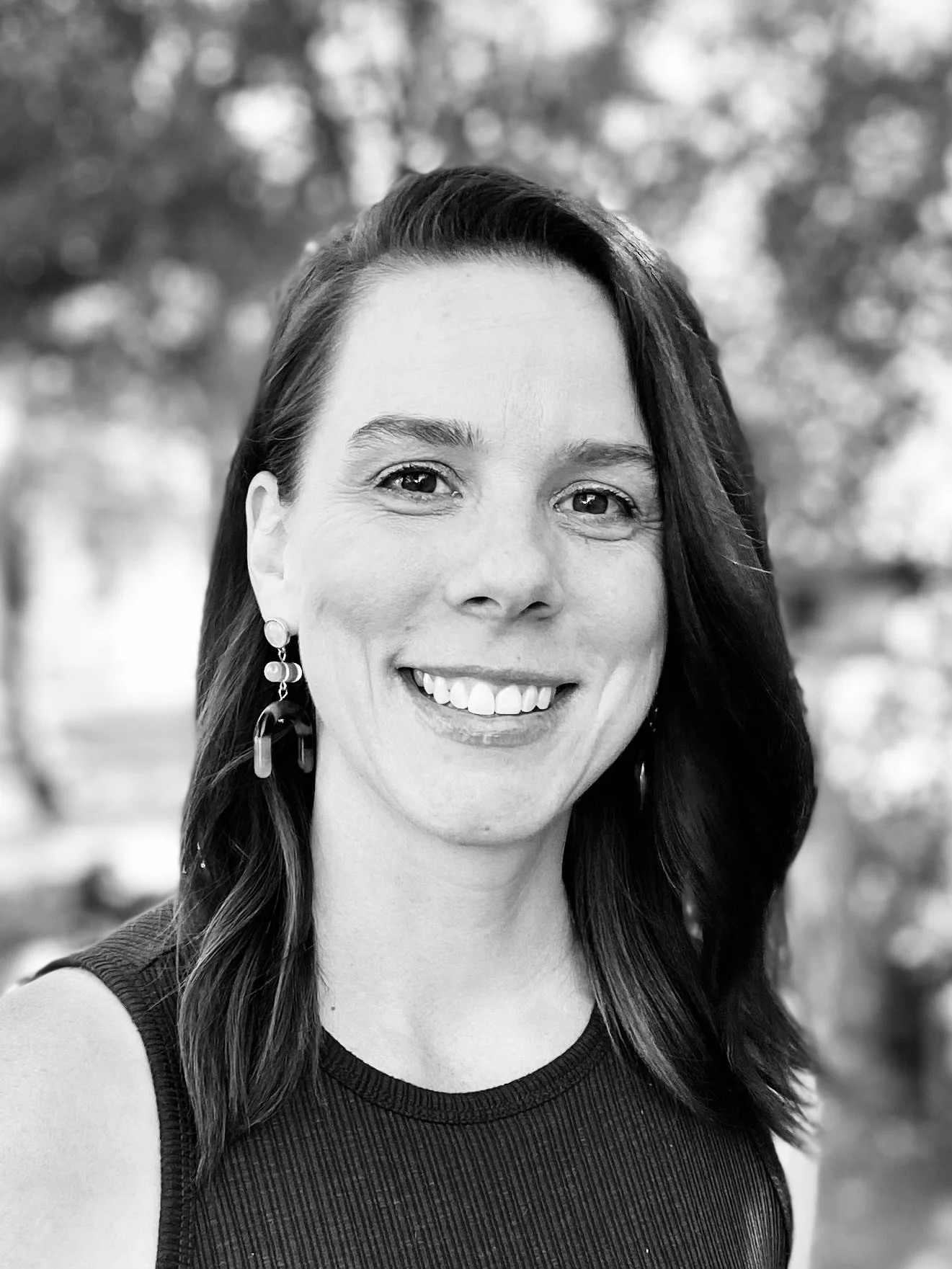 Black and white photo of a woman smiling outdoors, with blurred background trees. She has long dark hair, wears earrings, and a sleeveless top.