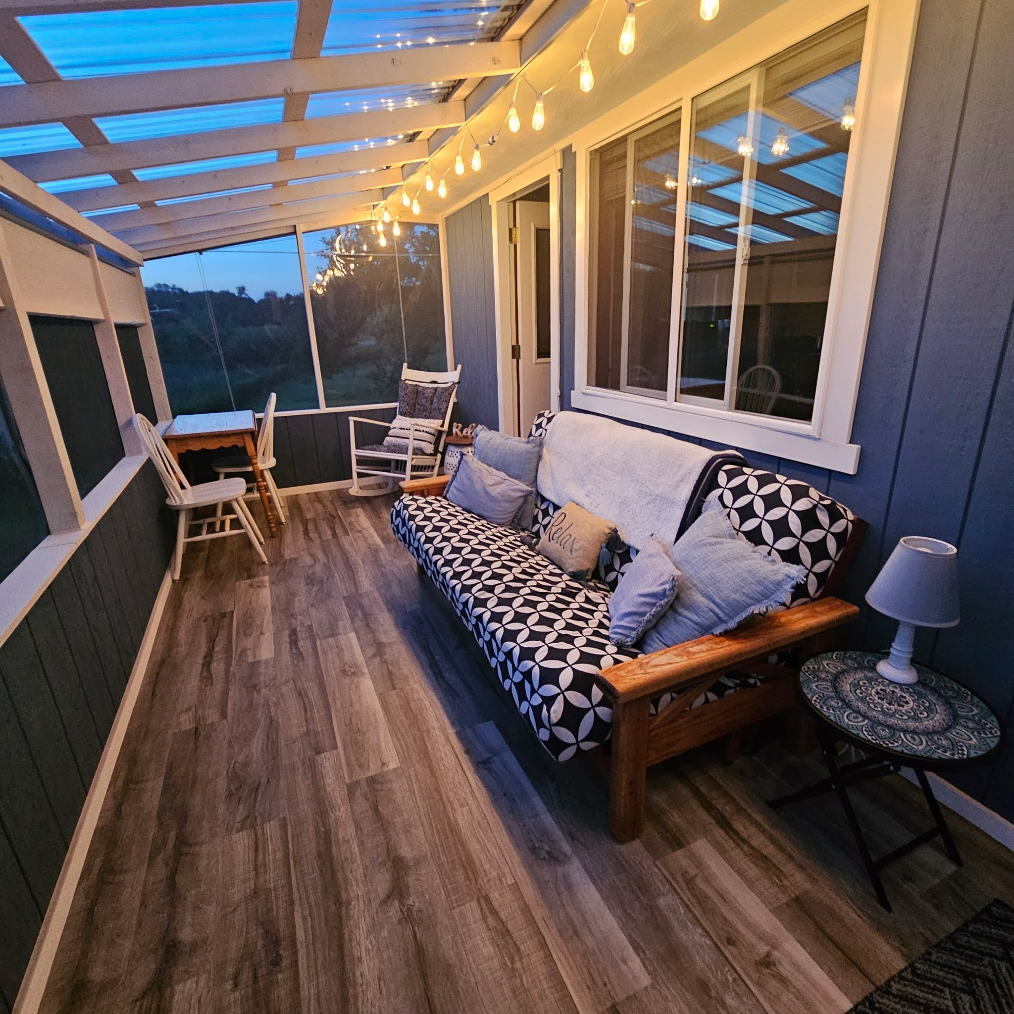 A cozy screened-in porch with string lights, a wooden floor, and a mix of cushioned seating and a small table, overlooking a view of trees at dusk.
