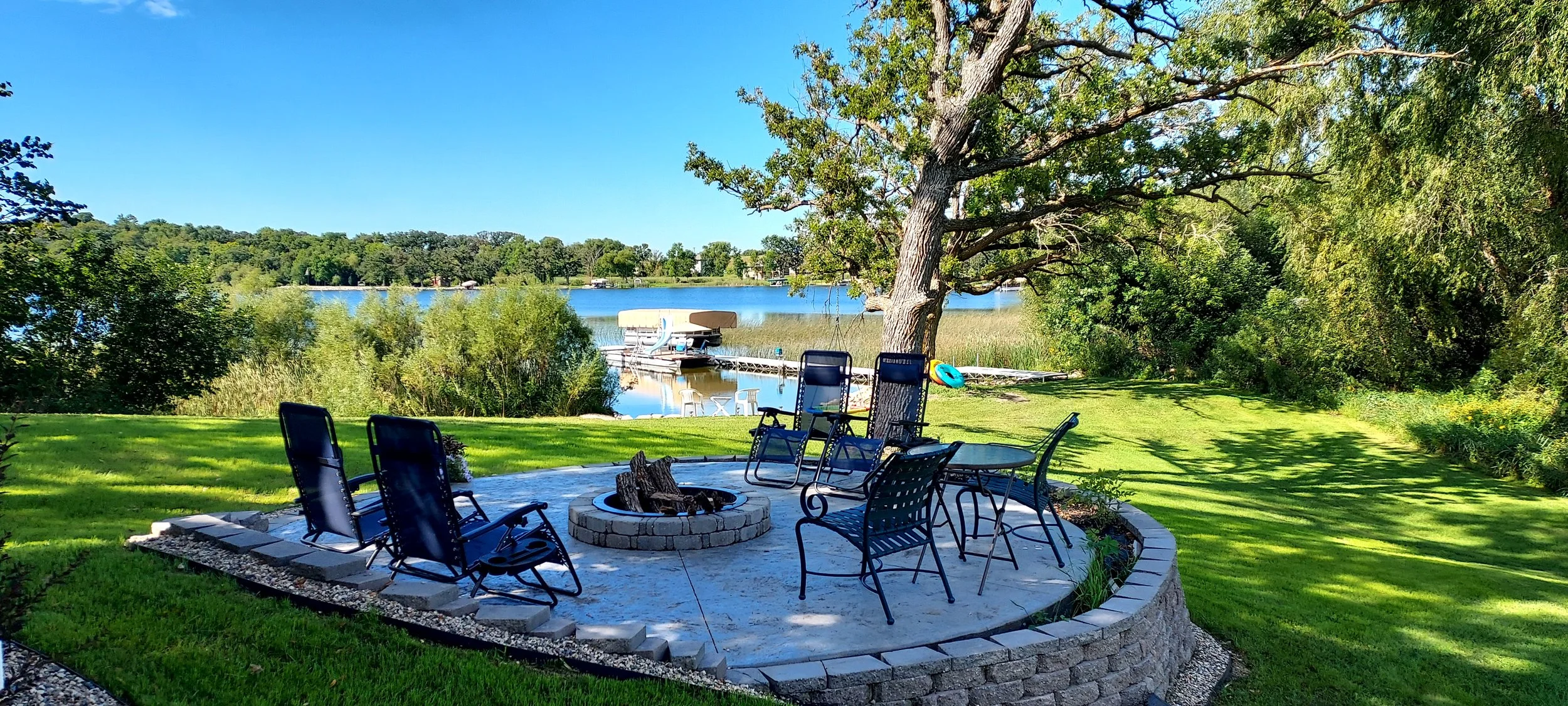 Lawn area with patio and chairs around a fire pit, next to a large tree with a lake in the background and boats docked on the water.