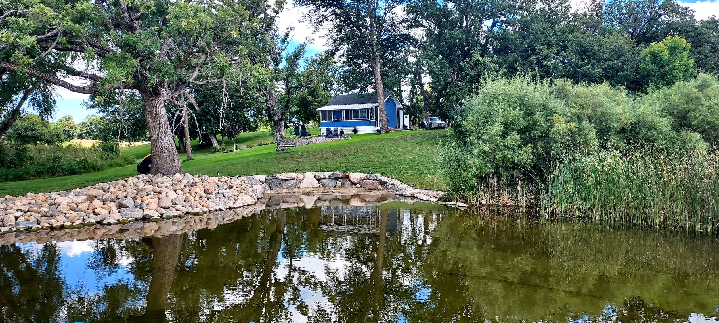 A small blue house with a porch is located on a grassy hill near a pond, surrounded by trees and bushes, with a rocky shoreline and some outdoor furniture.