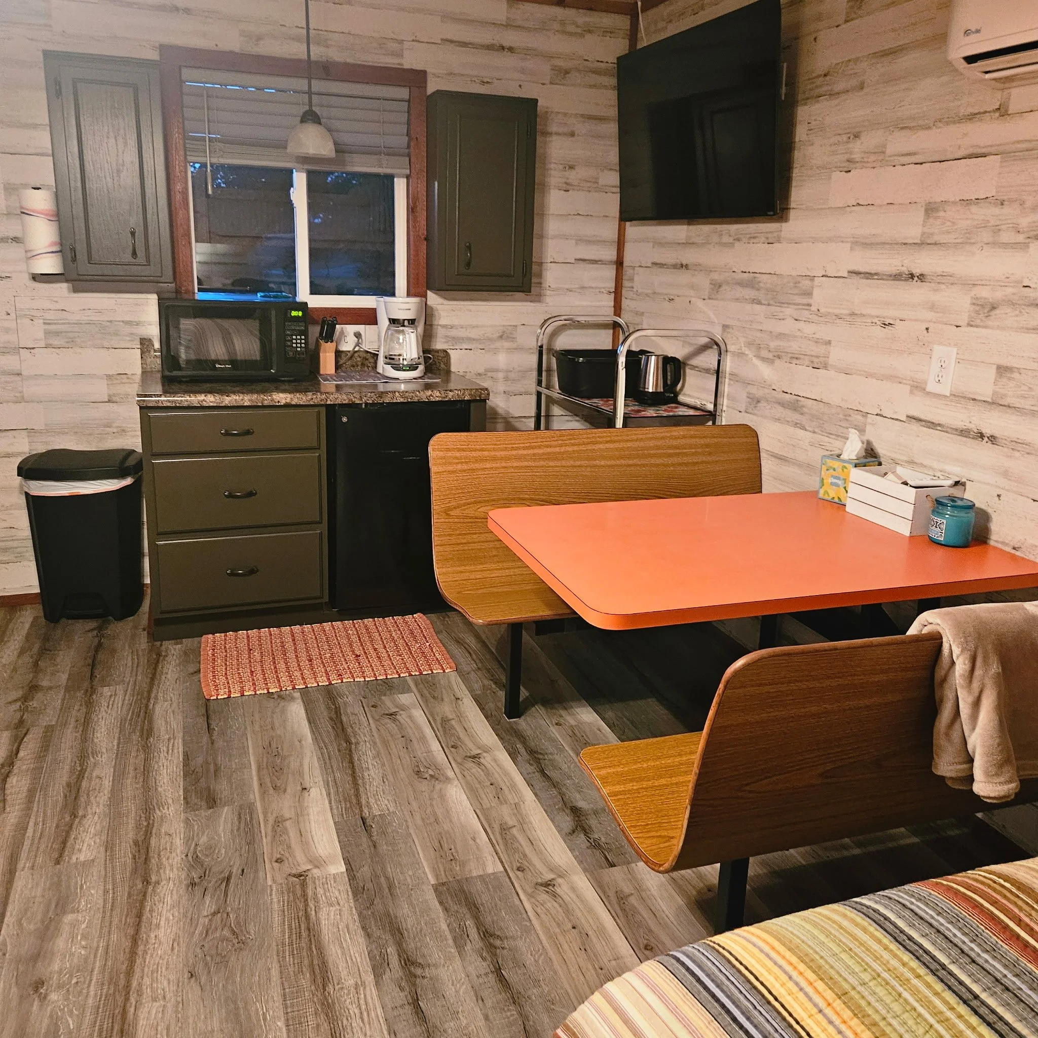 A cozy kitchen and dining area with a wooden wall, a small orange table with wooden chairs, a countertop with a microwave, coffee maker, and coffee pot, cabinets, a TV mounted on the wall, and various small items on the table and countertop.