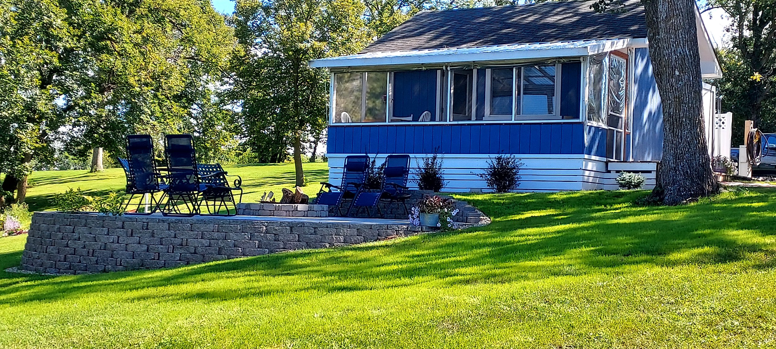 A blue and white house with a screened porch, green lawn, trees, garden chairs, and potted plants.