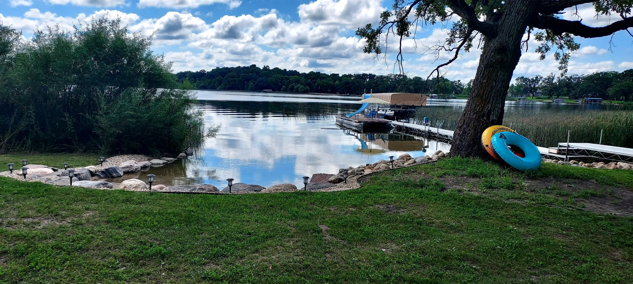 A lakeside scene with a grassy yard, a large tree, and two inflatable tubes leaning against the tree. The lake has a dock with watercraft and the sky is partly cloudy with reflections on the water.