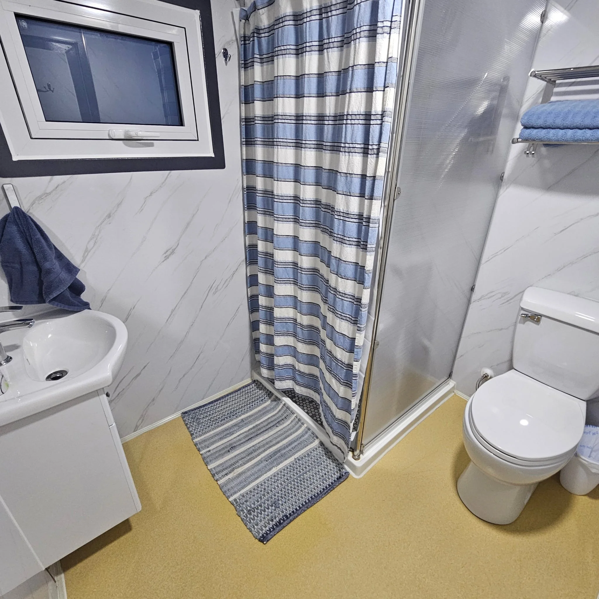 A small bathroom with a window, striped shower curtain, white toilet, small white sink, and a shelf with blue towels.