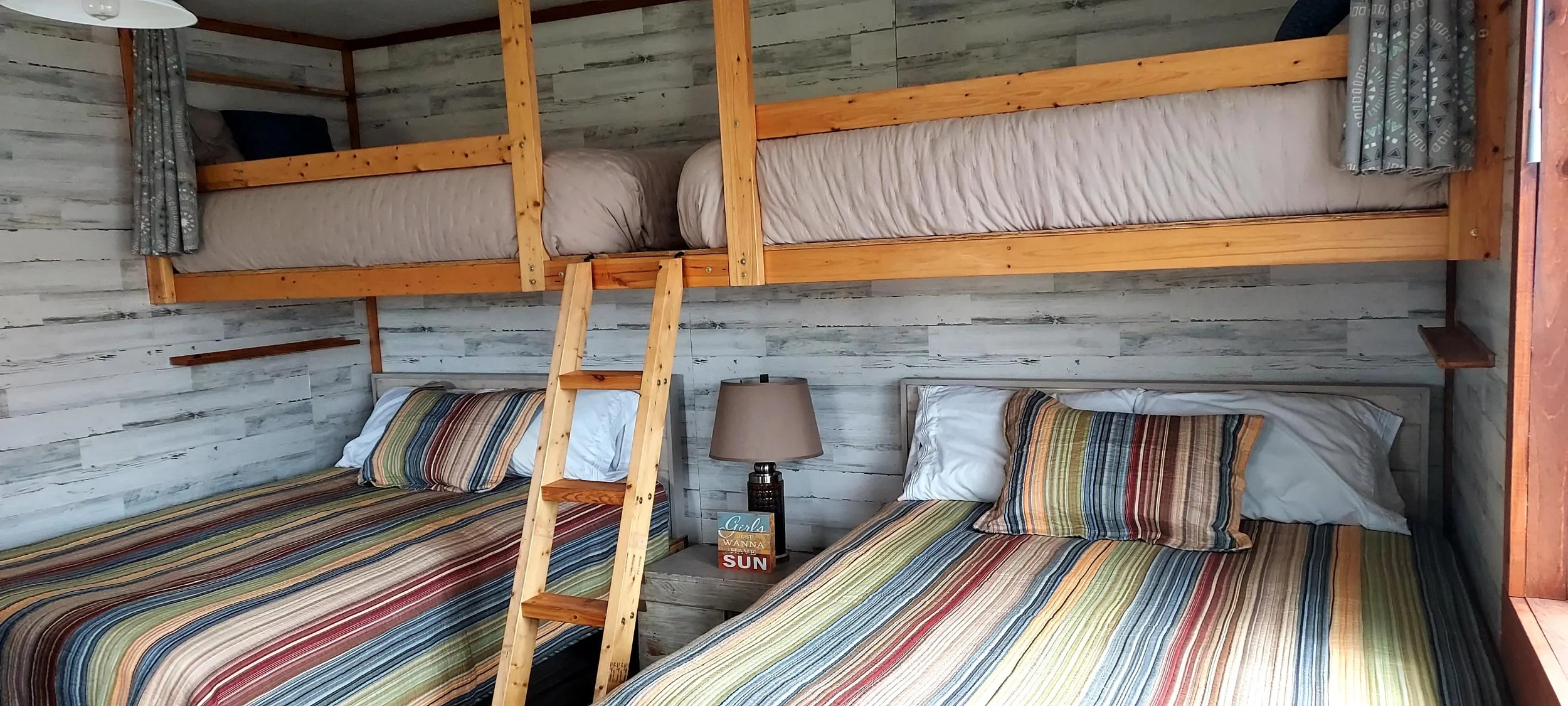 Bedroom with two beds, one lower and one upper, in a rustic cabin style. The lower beds have striped bedspreads with pillows, and the upper bed is accessed by a small wooden ladder. The walls are paneled with weathered white wood.