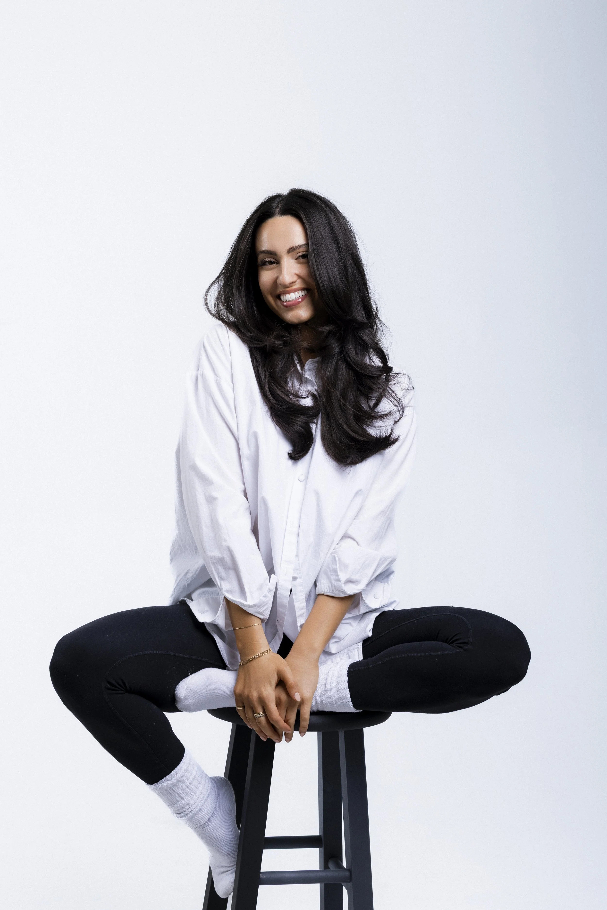 Woman in white shirt and black leggings sitting cross-legged on stool, smiling.