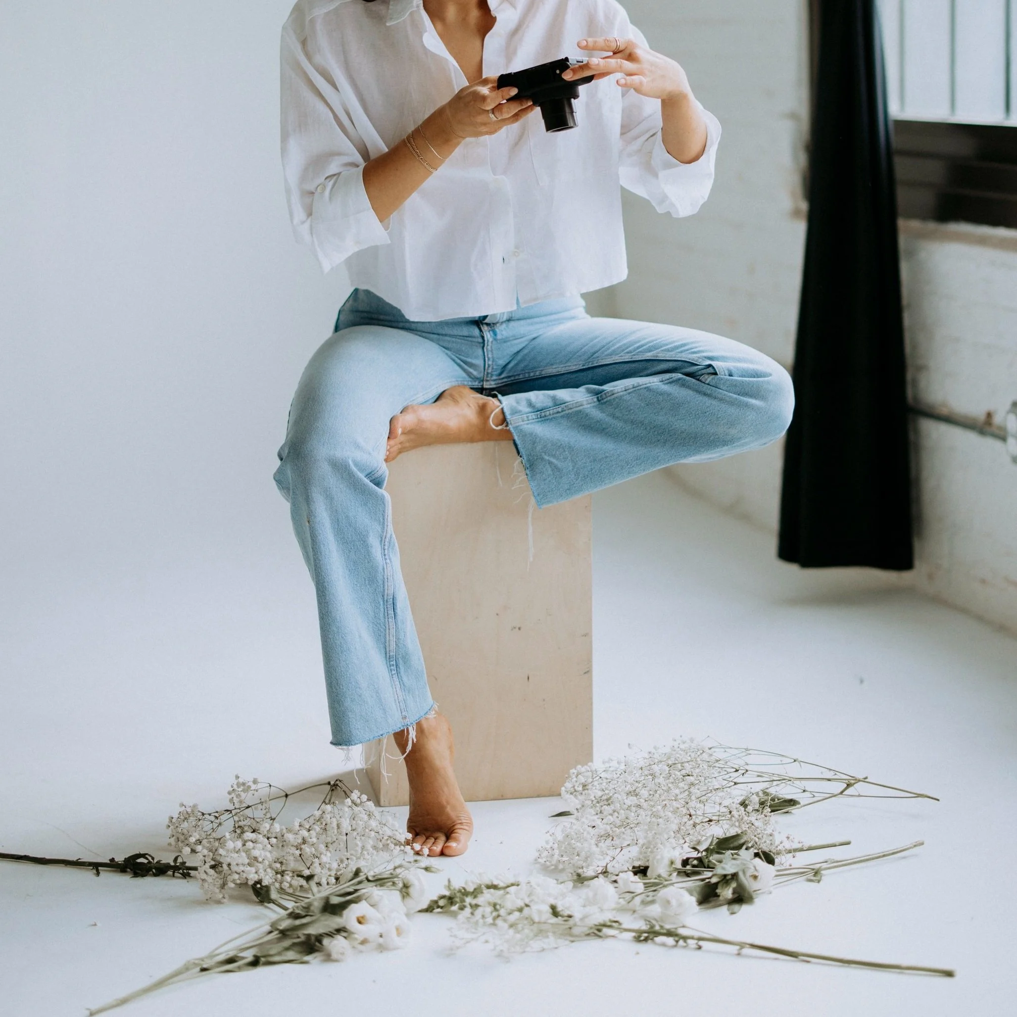 Person sitting on a box, wearing a white shirt and blue jeans, holding a camera, surrounded by white flowers on the floor.