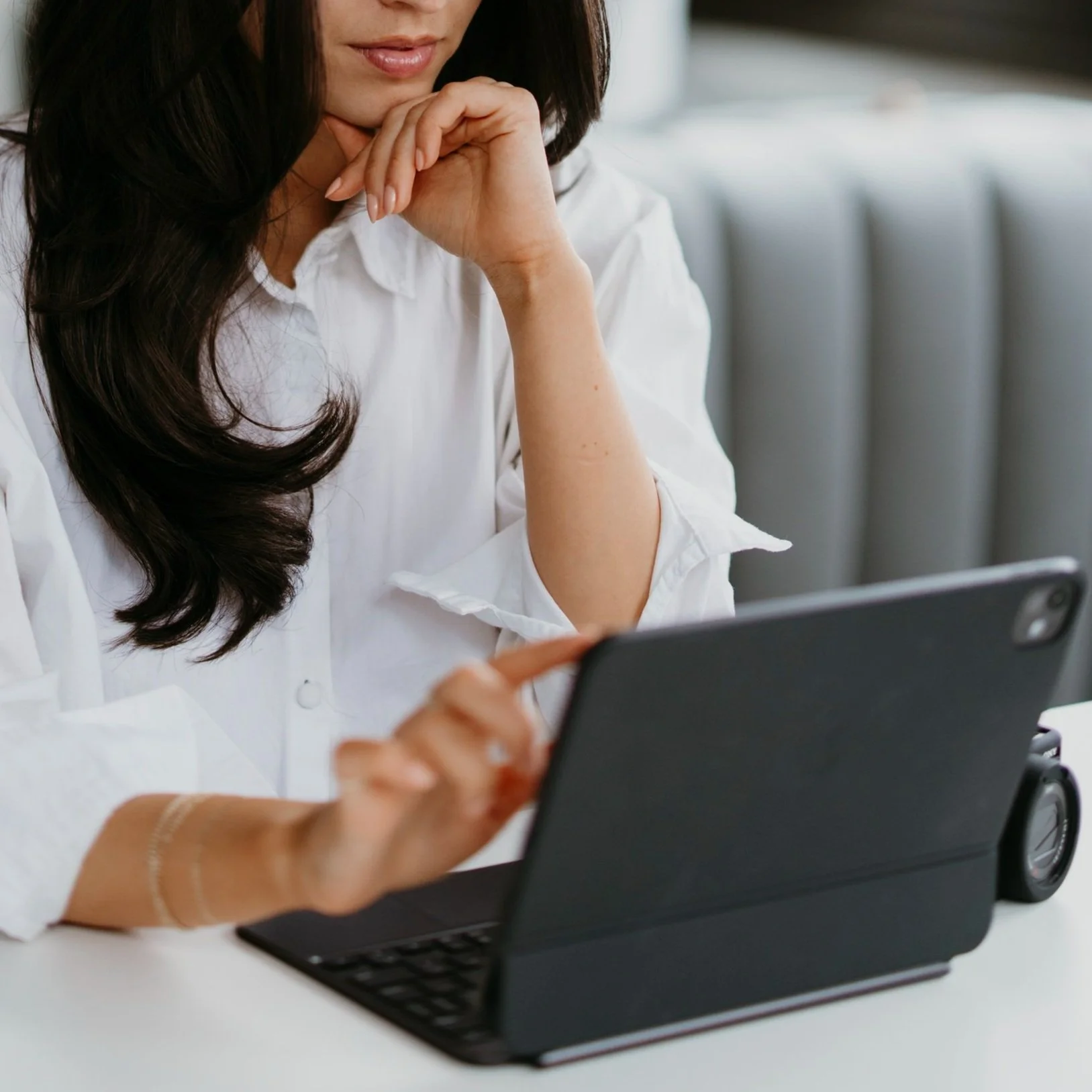 Woman using a tablet with a keyboard case, wearing a white blouse and seated in a modern setting.