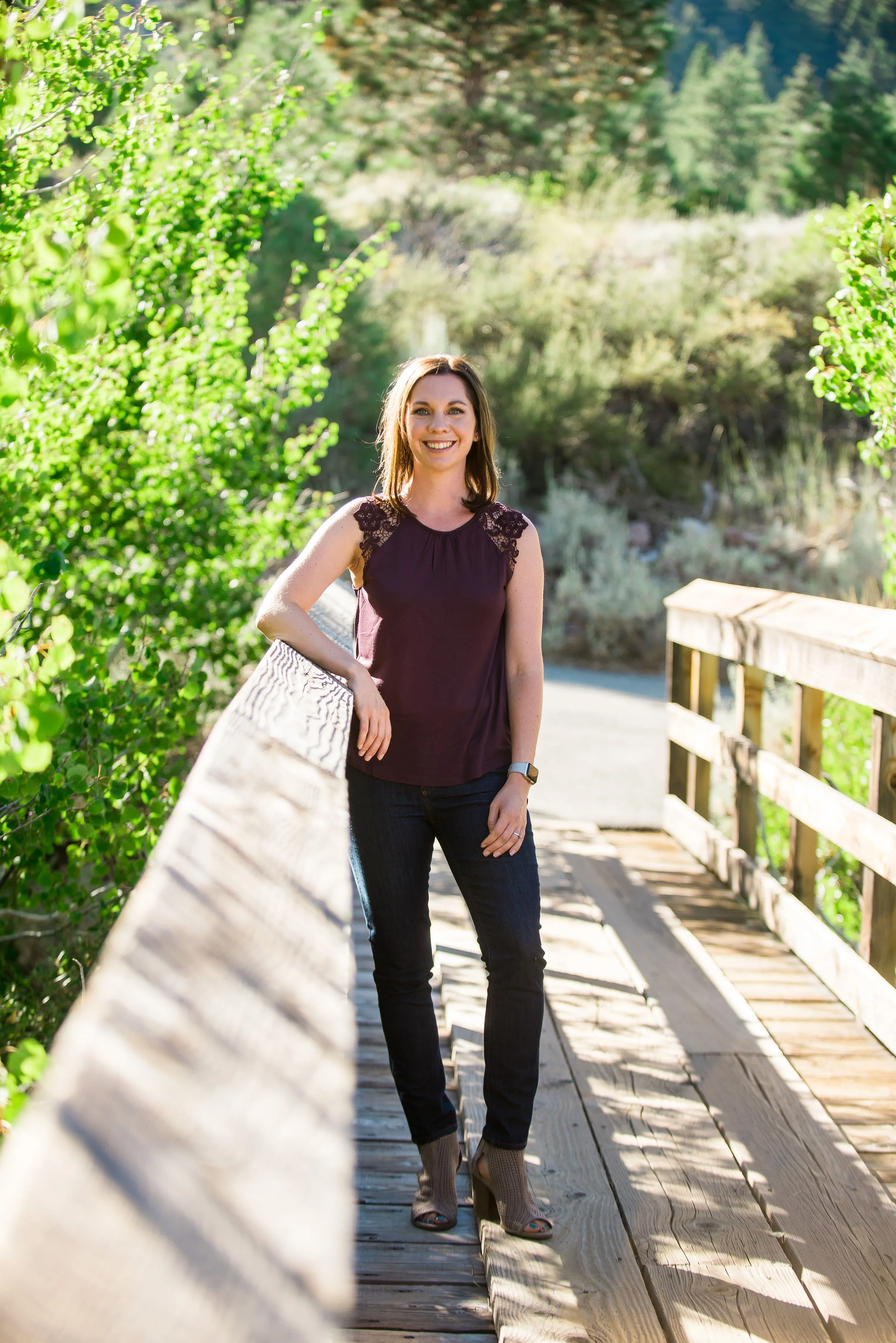 A woman with shoulder-length brown hair smiling, standing on a wooden bridge in a forested area, wearing a sleeveless maroon top, dark jeans, and beige high-heeled shoes.