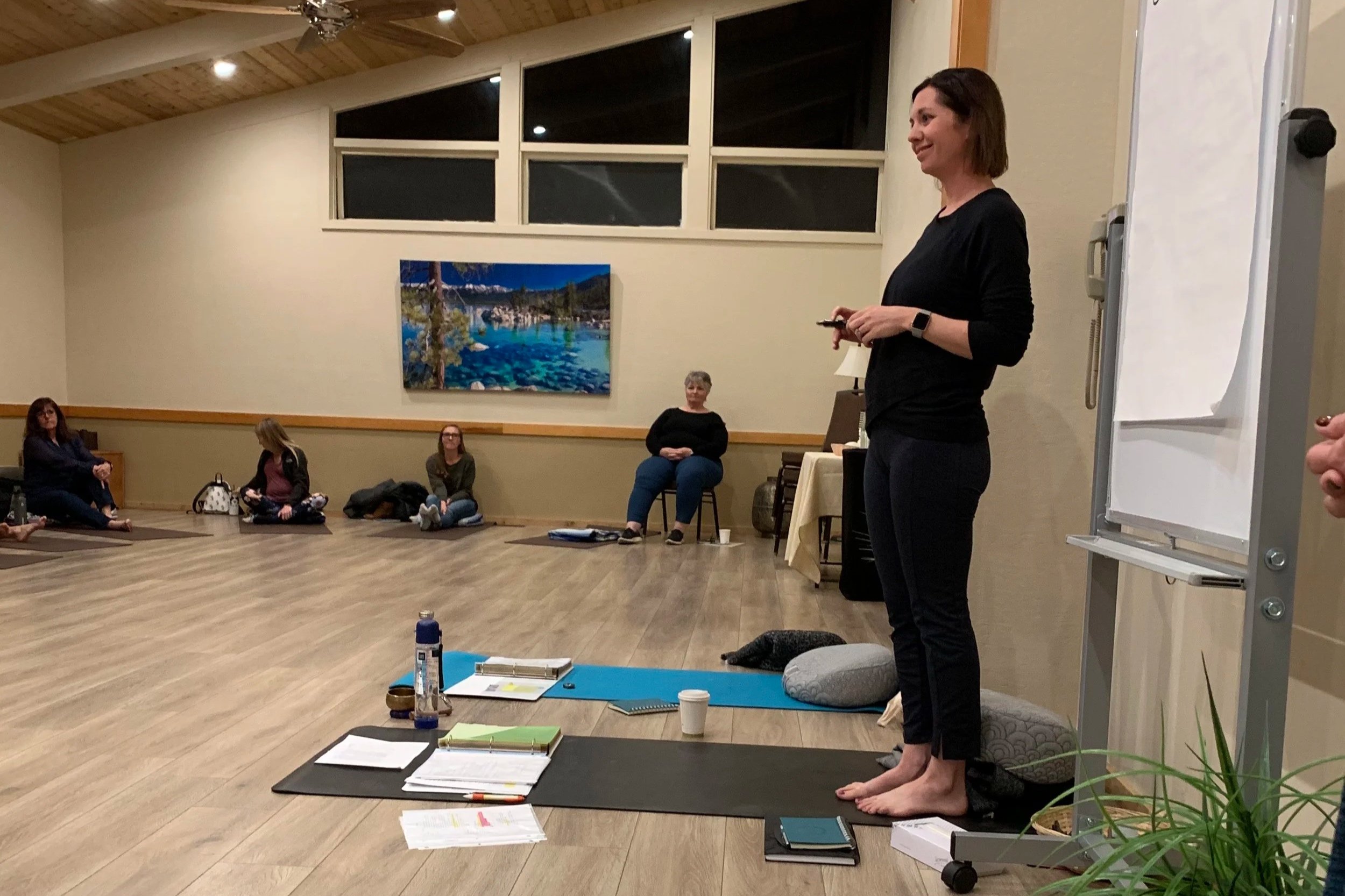 A woman standing barefoot on yoga mats in a room with high ceiling, holding a phone, leading a group discussion or meditation session, with seated participants across the room, a whiteboard to her right, and a large landscape painting on the wall.