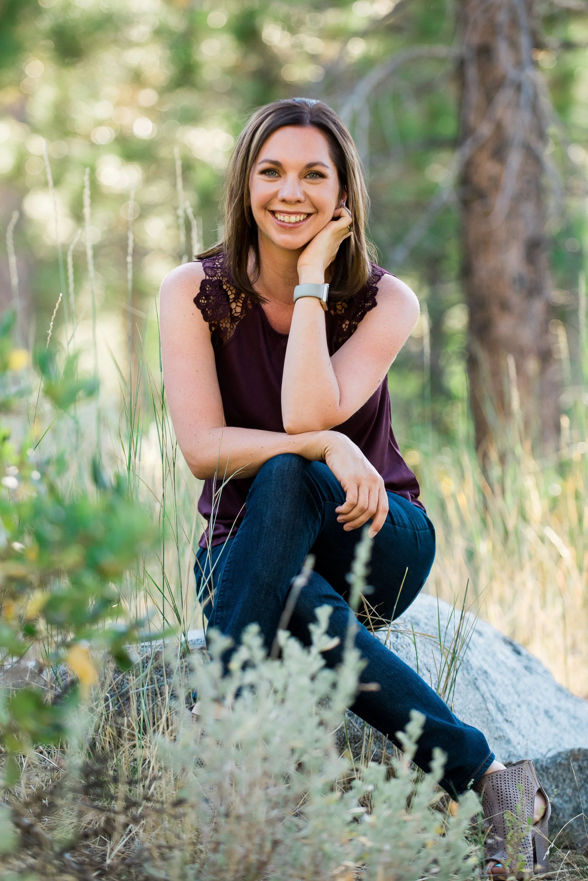 A woman with shoulder-length brown hair, wearing a sleeveless purple top with lace details on the shoulders, jeans, and open-toed shoes, sitting on a rock in a forested area with greenery and trees in the background, smiling at the camera.