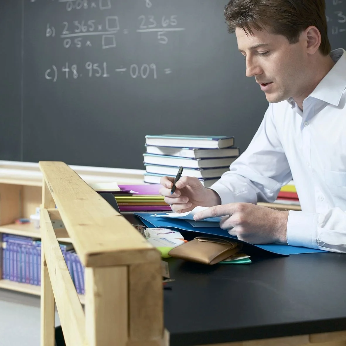A teacher in a white shirt sits at a desk grading papers.