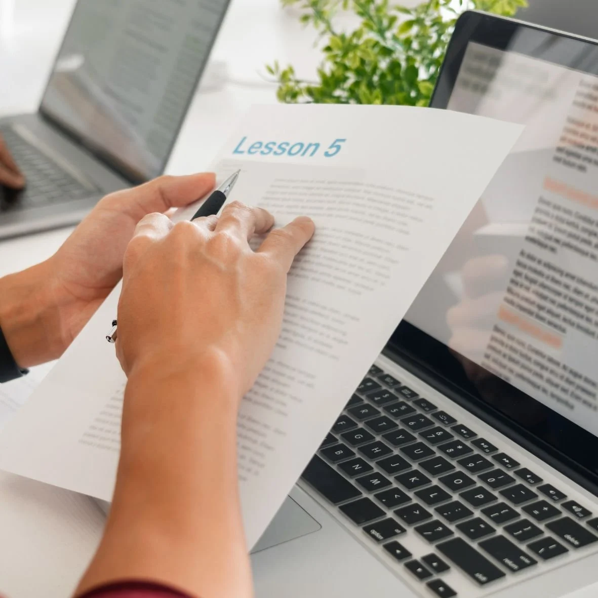A pair of hands hold a pen and a packet of papers in front of a laptop screen.