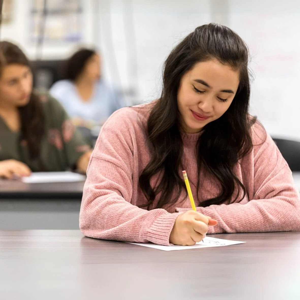 A student with a pink shirt writes on a sheet of paper with a pencil.