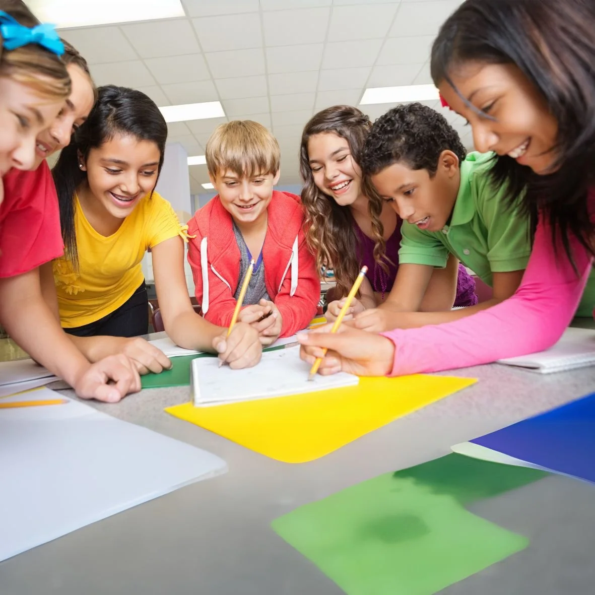Photo of six students and a teacher gathered around a sheet of yellow paper on a table.