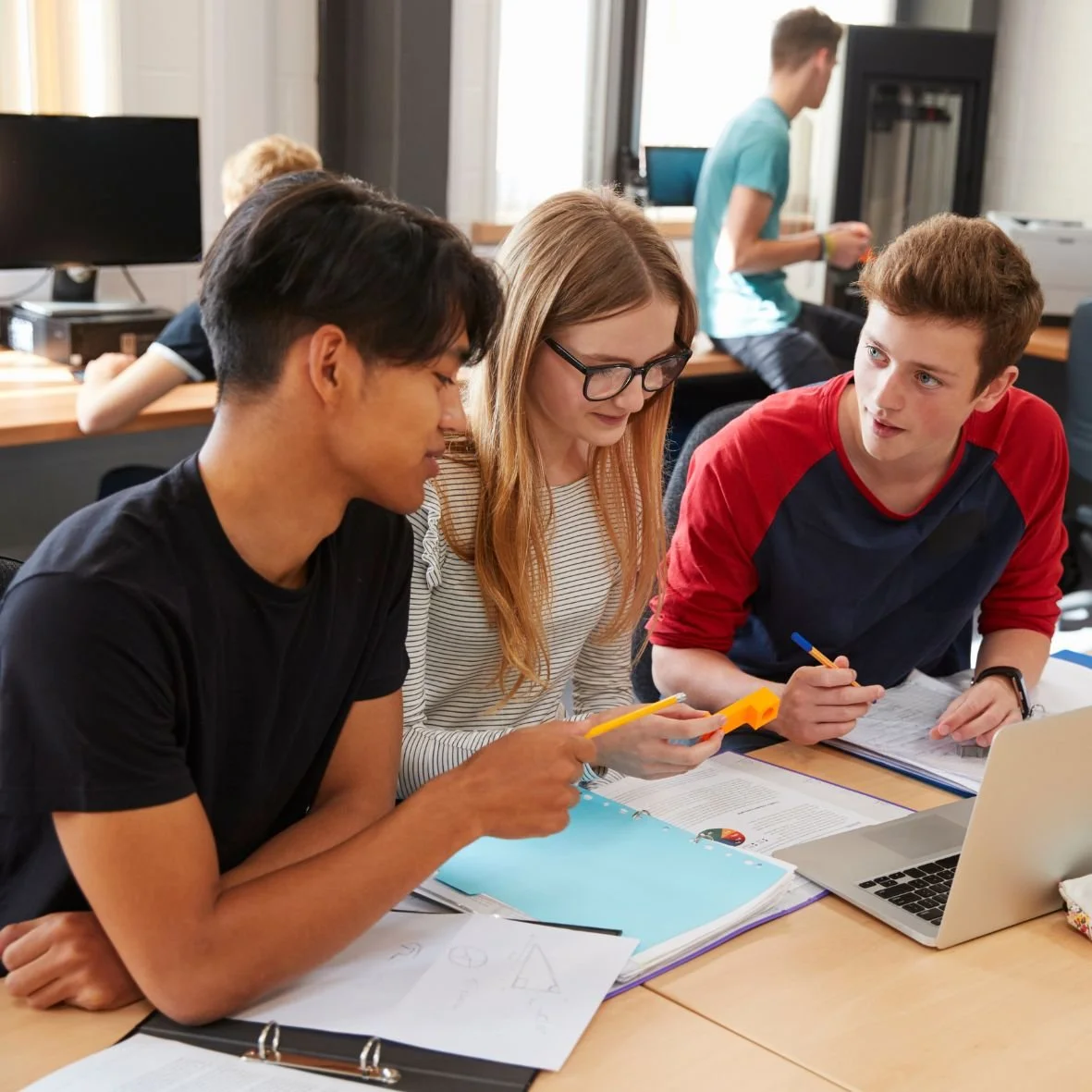 Three teenage students sit at a lab table discussing an assignment.
