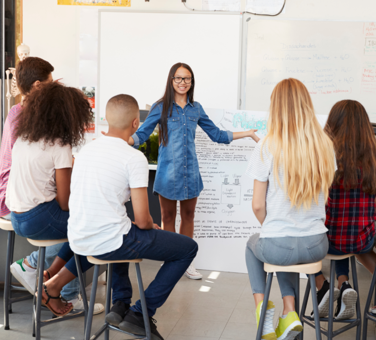 Photo of a teacher pointing at a whiteboard, standing in front of five teenage students who are seated on stools.