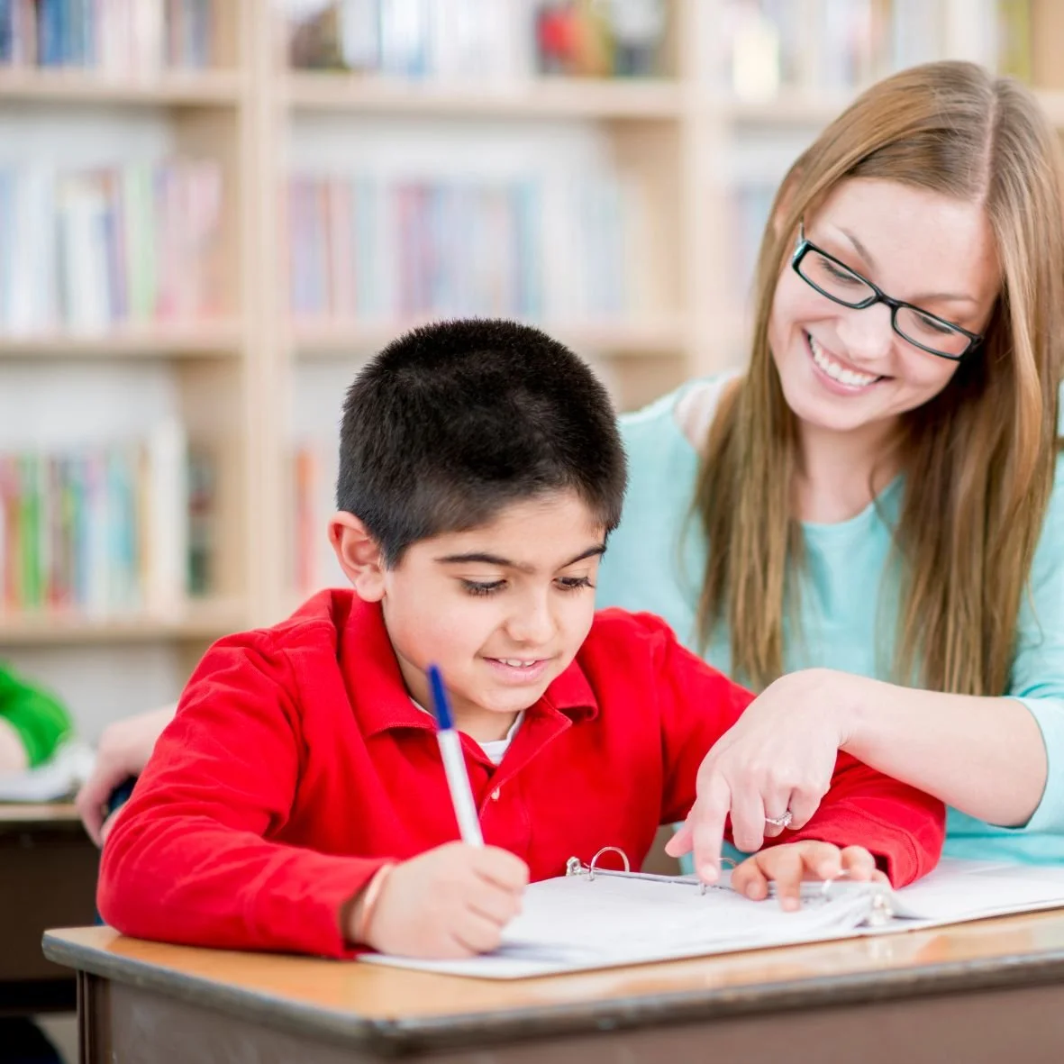 A smiling teacher stands over the shoulder of a student in a red shirt.