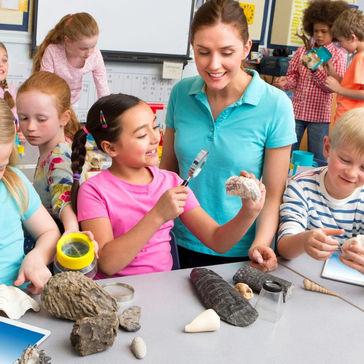 A smiling teacher in a blue shirt stands with a student in a pink shirt, looking through a magnifying glass.