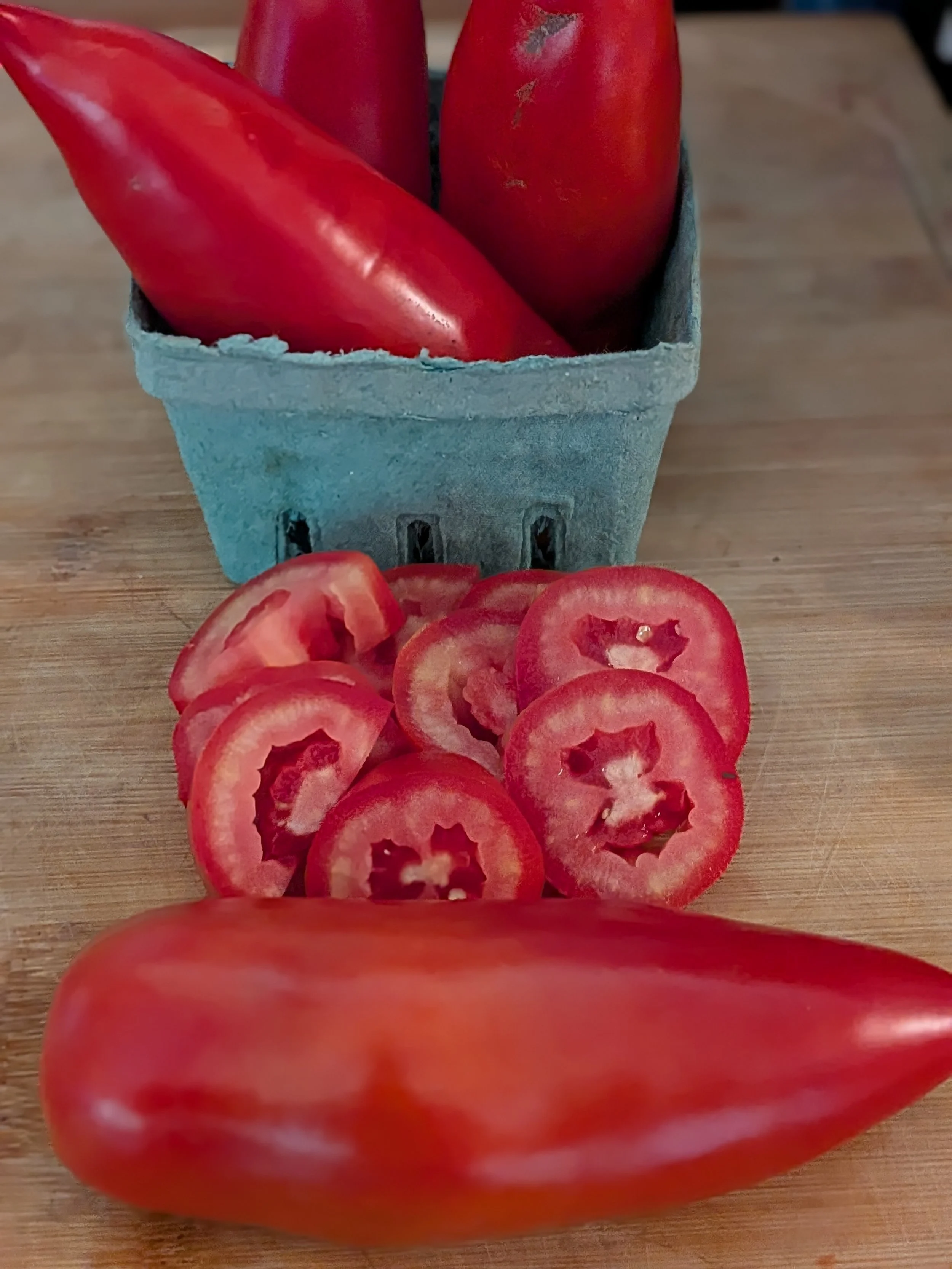 A red heirloom tomato and sliced tomato rounds on a wooden cutting board, with a gray egg carton holding more tomatoes in the background.