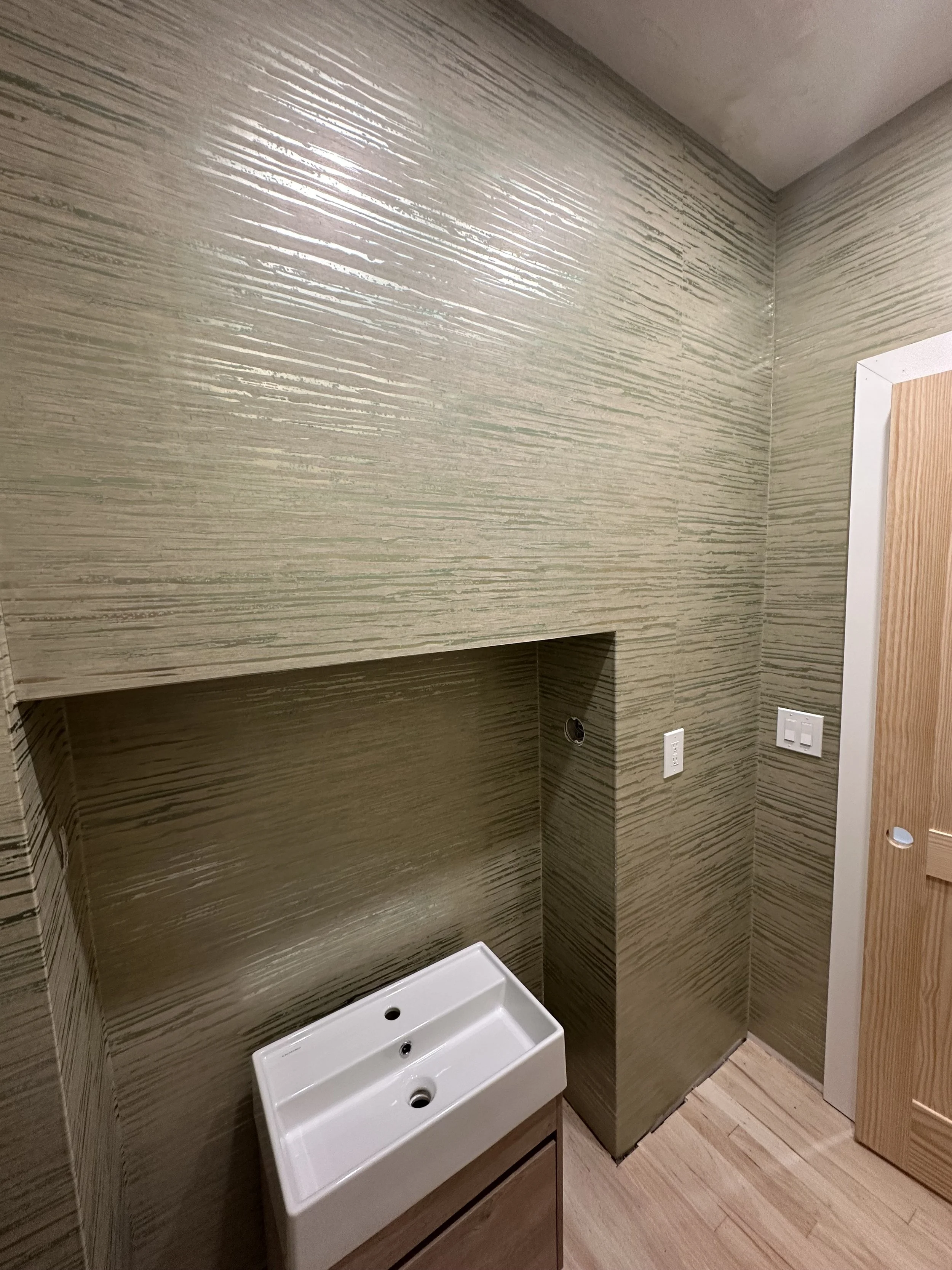 A partially installed bathroom corner with textured beige wallpaper, a small white sink, a wall outlet, and a wooden door.