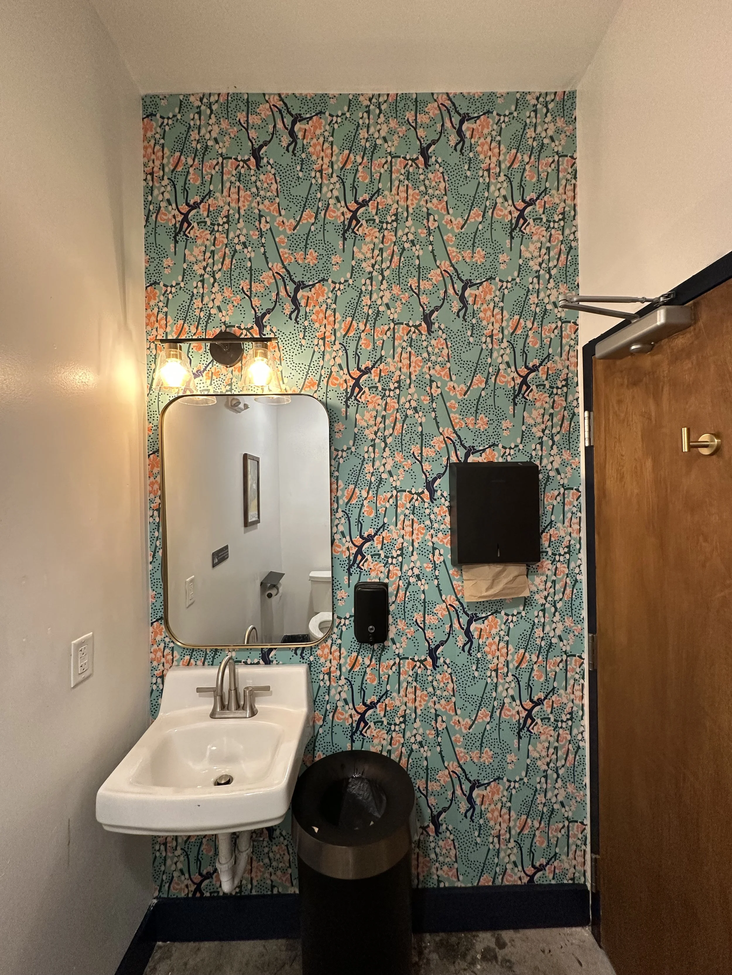 Small bathroom with a floral patterned accent wall, a mirror above a small white sink, a trash can, and a wooden door.