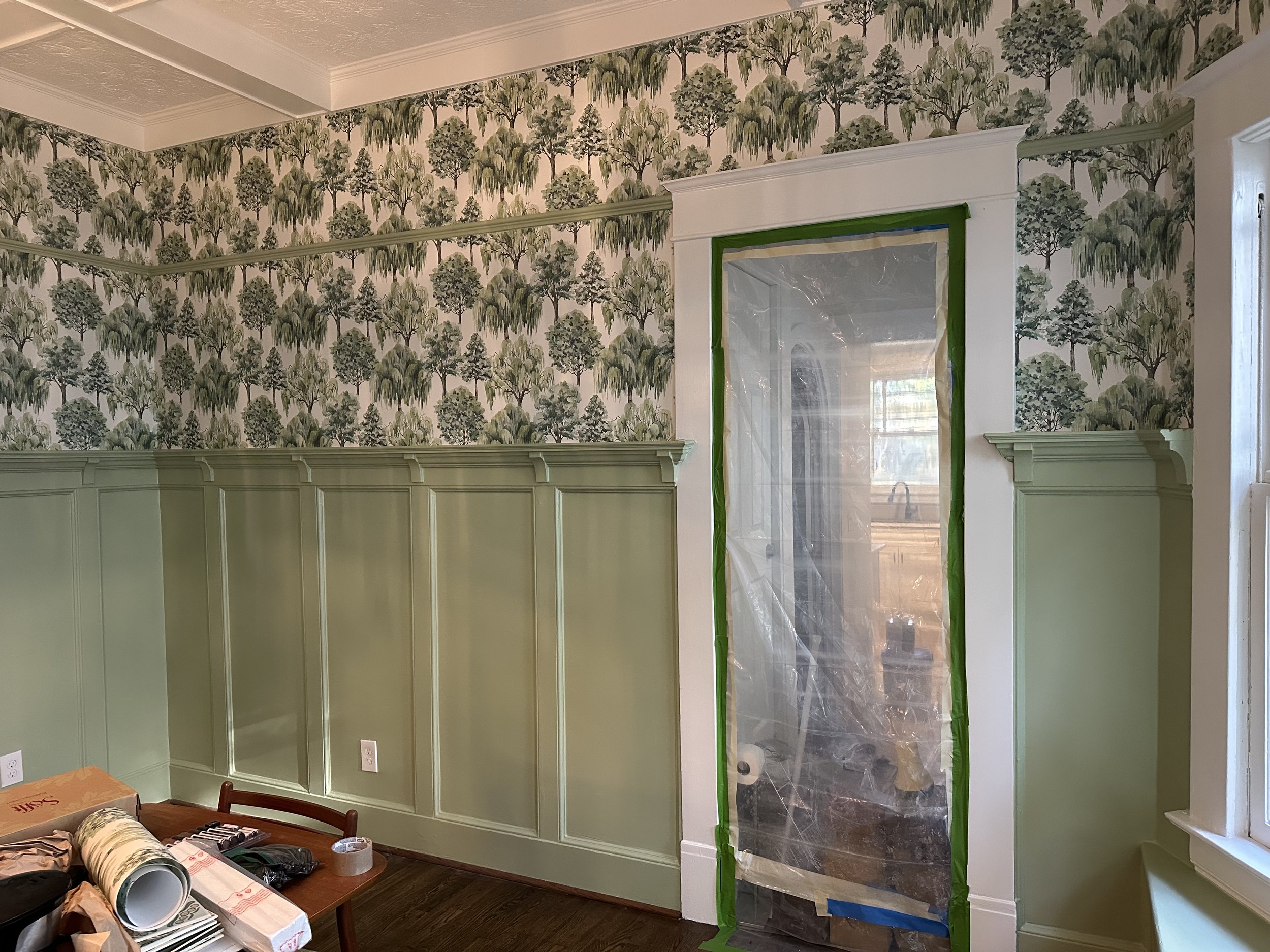 Interior room with green and white wainscoting, wallpaper with trees, and a doorway covered with plastic sheeting and painter's tape, indicating ongoing painting or renovation work.