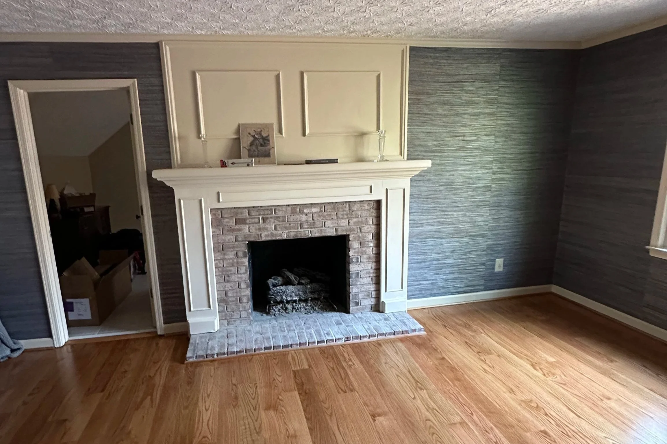 Empty living room with hardwood floors, a brick fireplace with white mantel, and gray textured wallpaper on walls. An open door reveals a room with boxes and furniture.