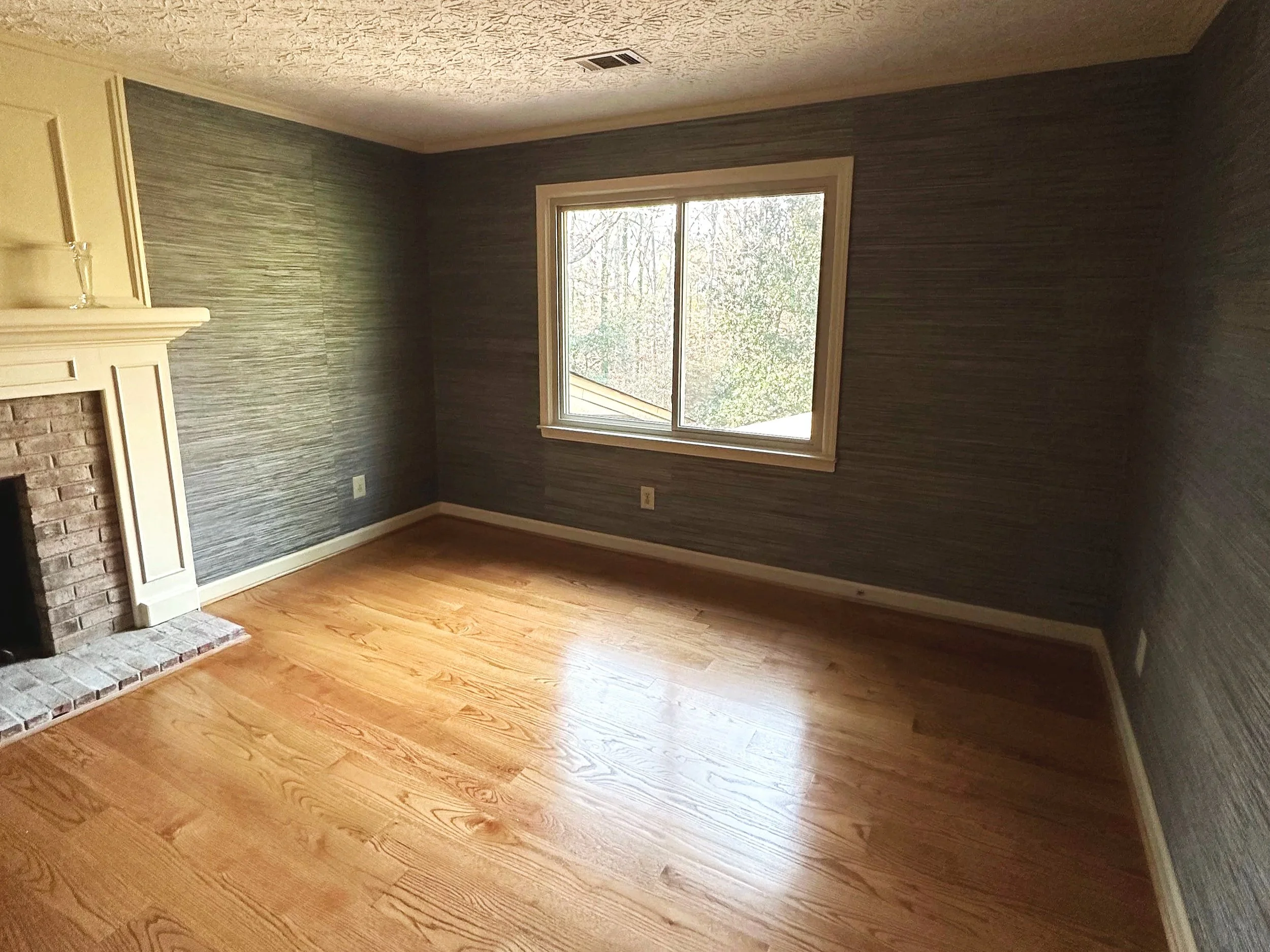 Empty room with wooden flooring, dark textured wallpaper on the walls, a window with white trim, and a brick fireplace with a white mantel.
