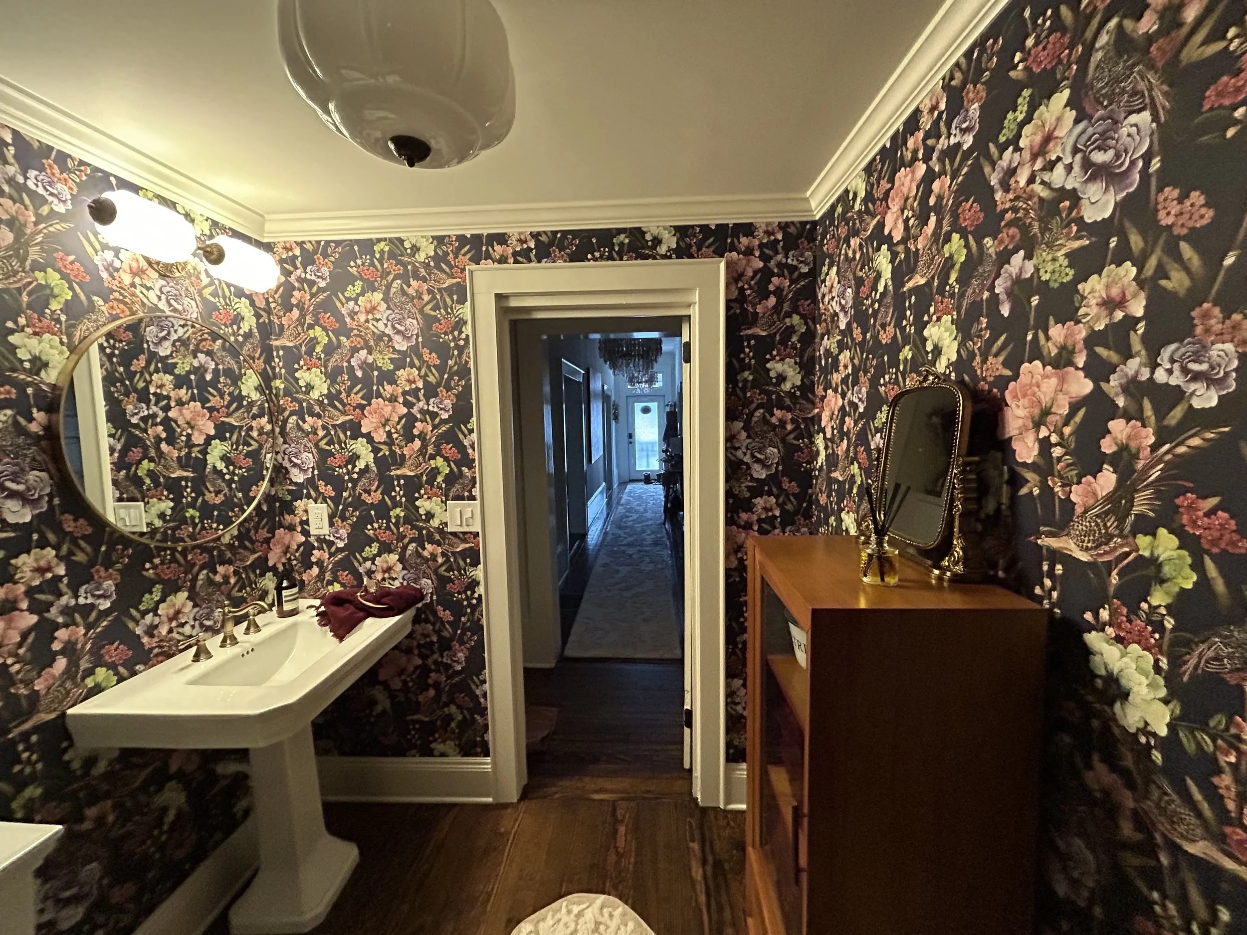 Bathroom with floral wallpaper, a white sink, a mirror, and a wooden shelf with a small mirror and decorative items.