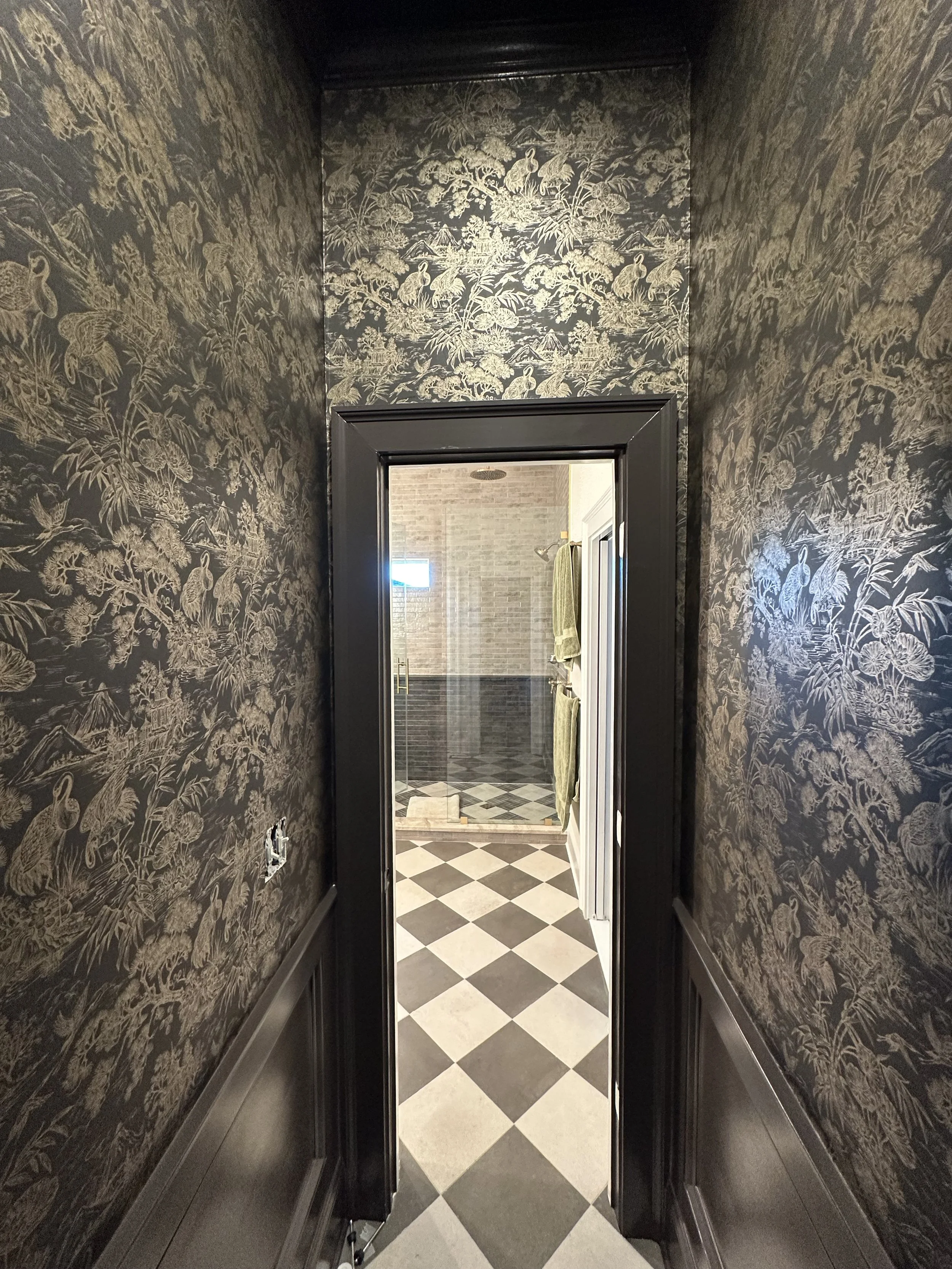 View down a narrow black hallway with intricate wallpaper featuring cranes, leading to a bathroom with checkered black and white tile flooring, a glass shower, and a towel hanging on the wall.